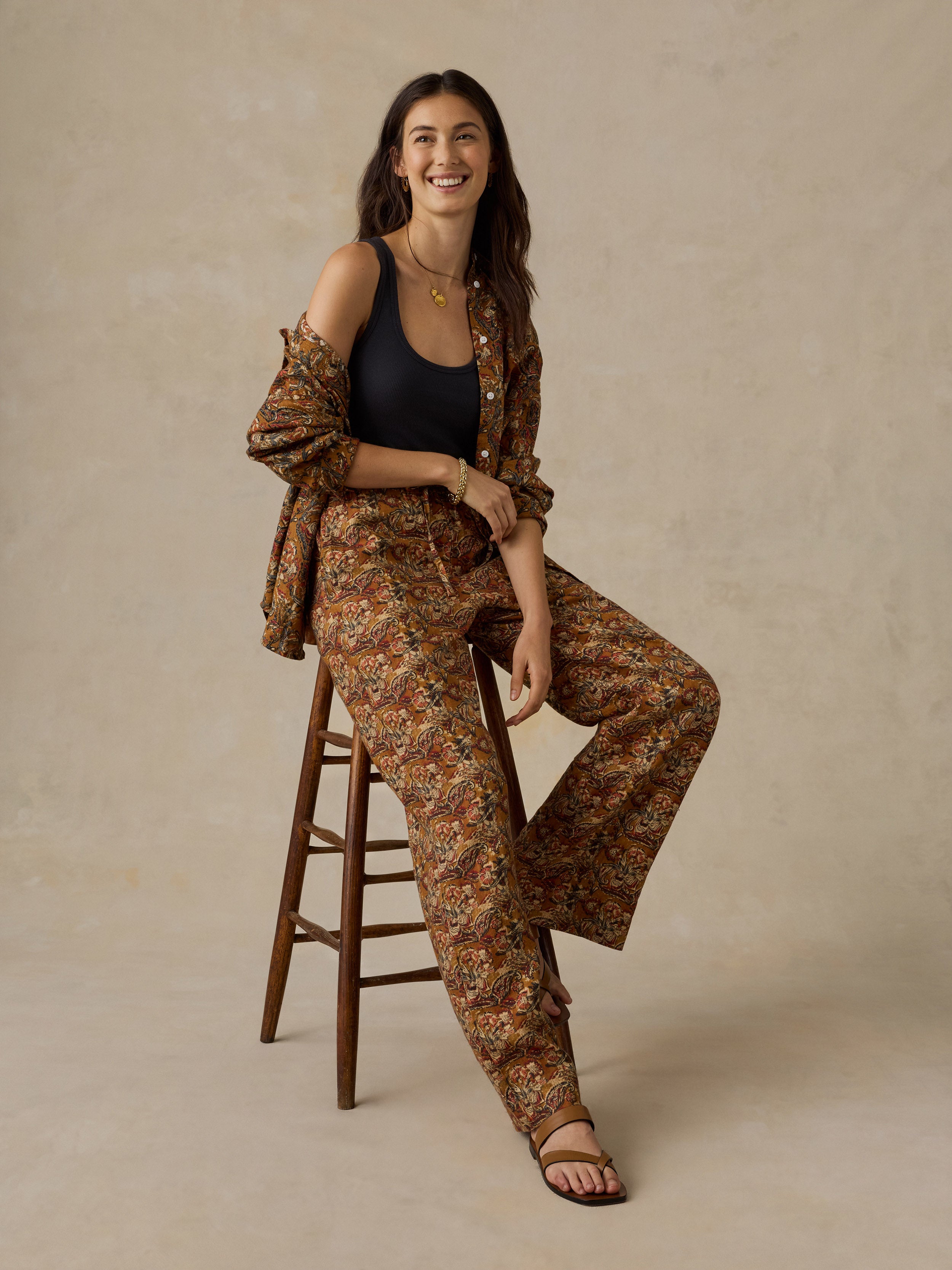 Woman sitting on a wooden stool wearing an orange floral printed button up shirt and matching pants against a beige background