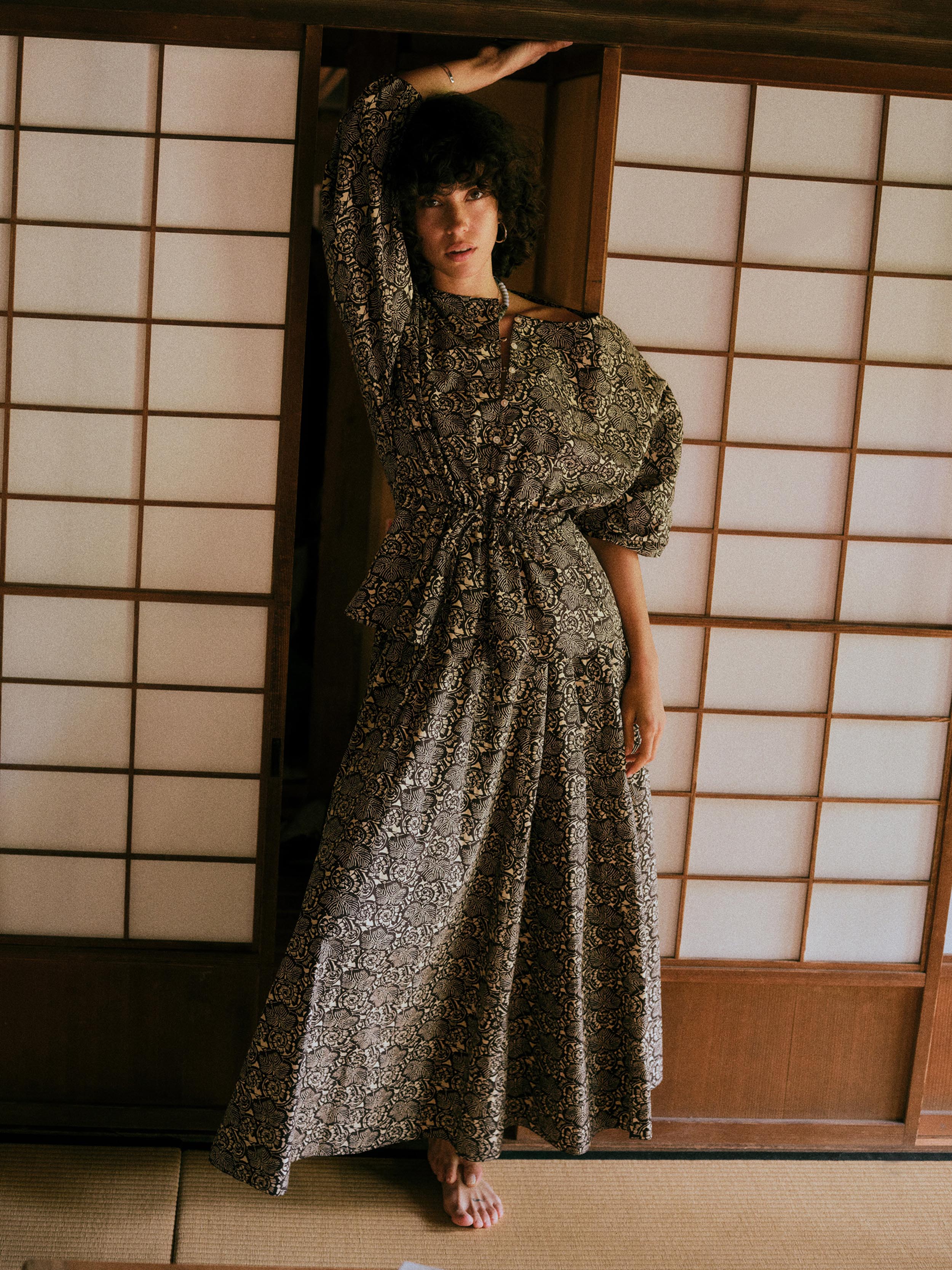 A woman wearing a long, loose-fitting floral top and matching maxi skirt with a dense beige and black floral pattern, posing in front of traditional Japanese shoji screens.