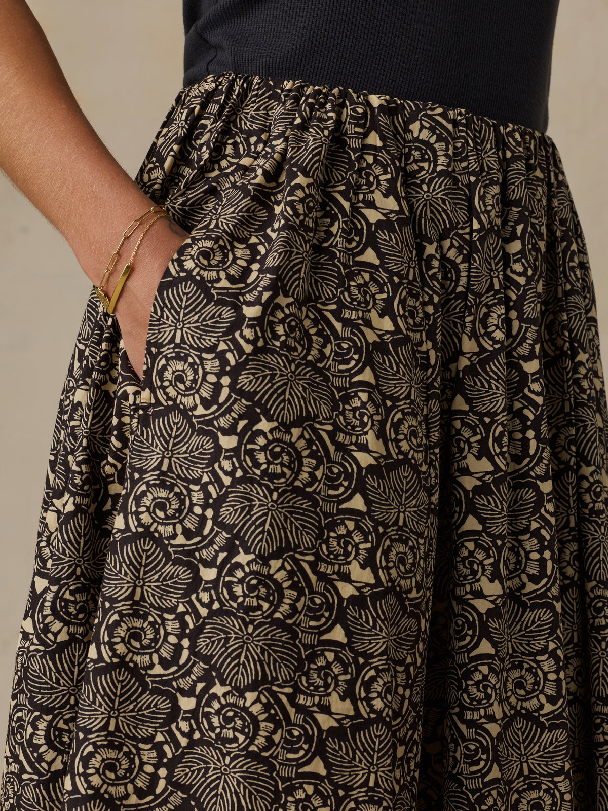 Close-up of a person wearing a black and beige floral print skirt with side pockets, paired with a dark navy top. The person's hand is in the pocket, and they are wearing gold bracelets., image 6 of 6