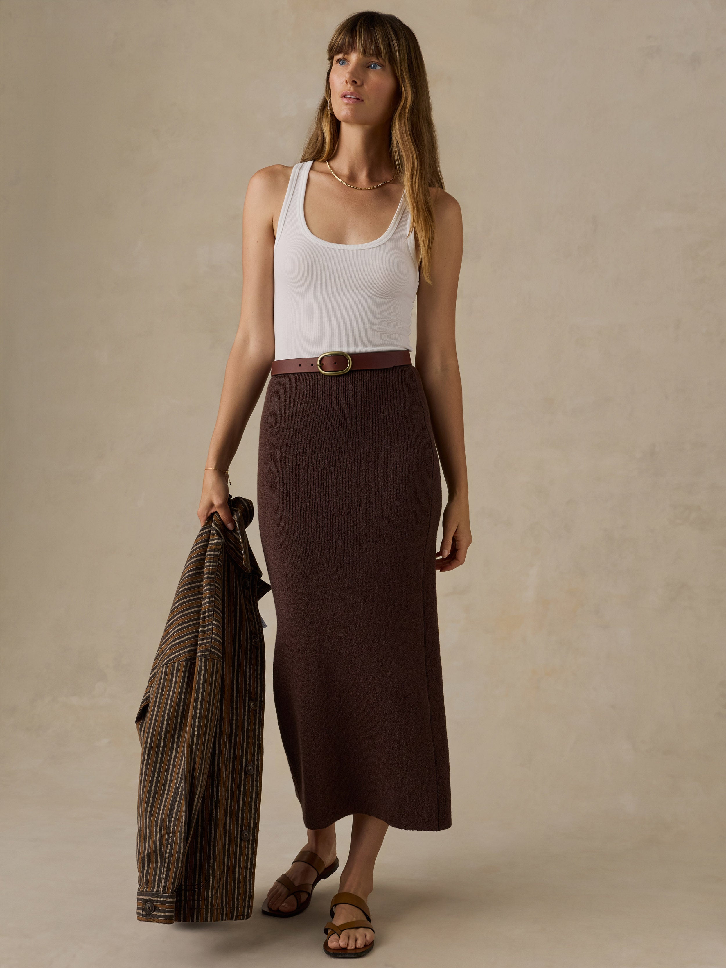Woman wearing a white tank top and brown knit skirt holding a brown striped jacket against a beige background