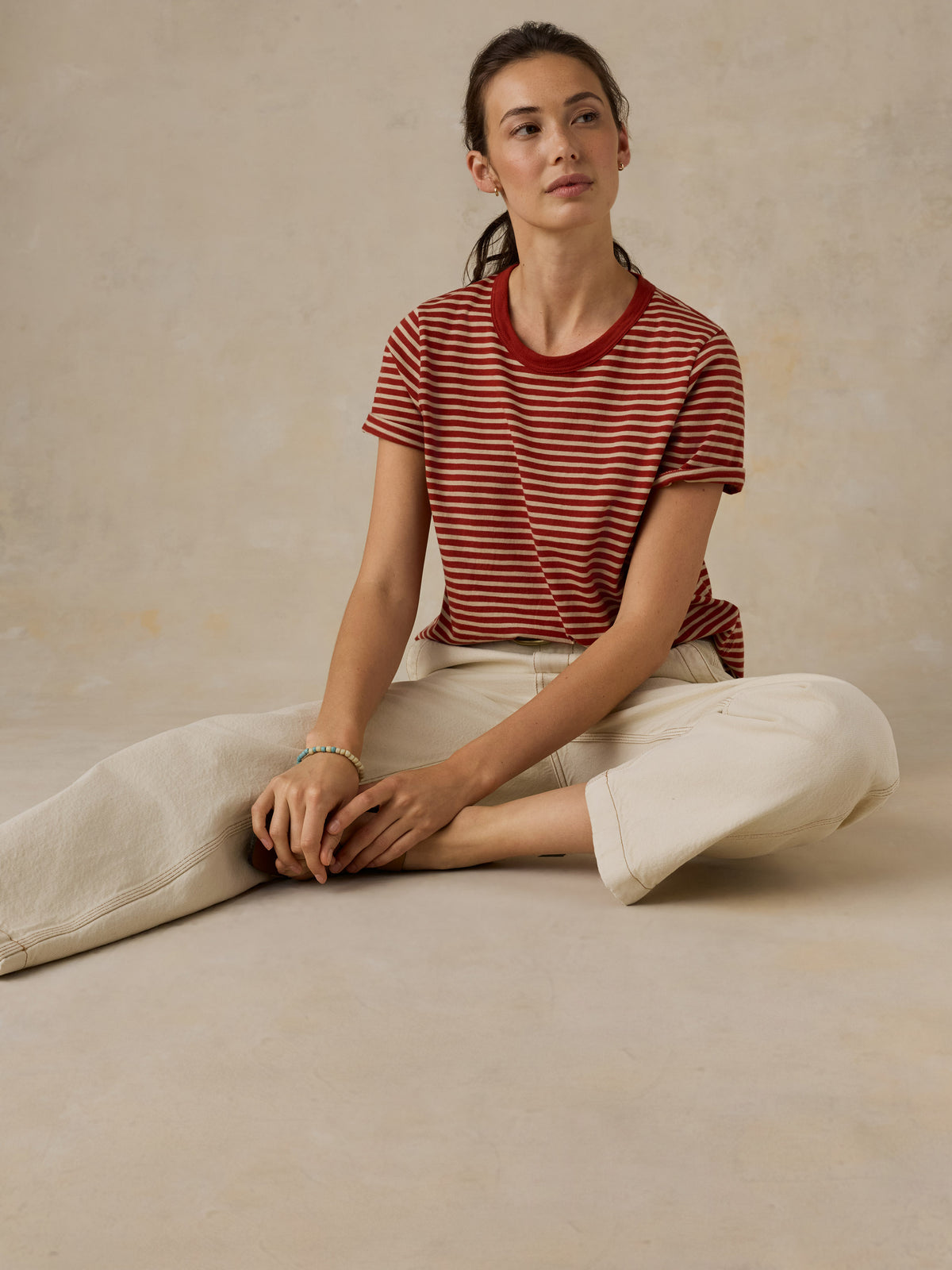 Woman sitting on the ground wearing a red and white striped t-shirt and cream colored denim jeans against a beige background, image 4 of 7
