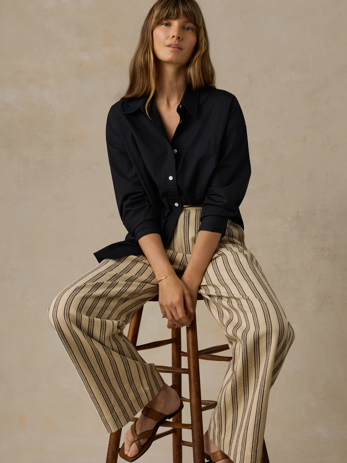 Woman sitting on a wooden stool wearing a black button down shirt and beige and black striped pants against a beige background. , image 3 of 6