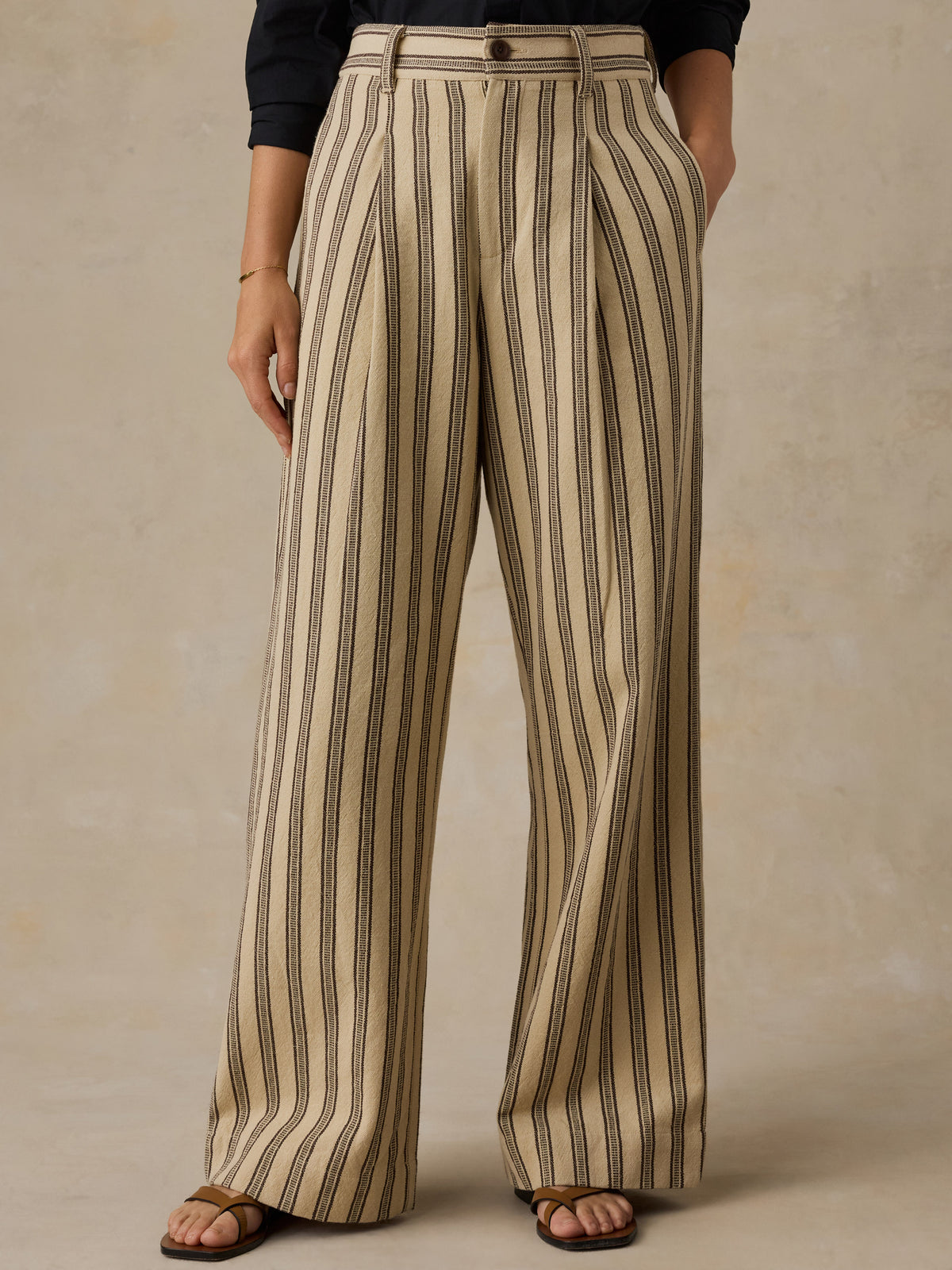 Woman wearing a black button down shirt and high waisted beige and black striped pants against a beige background. , image 2 of 6