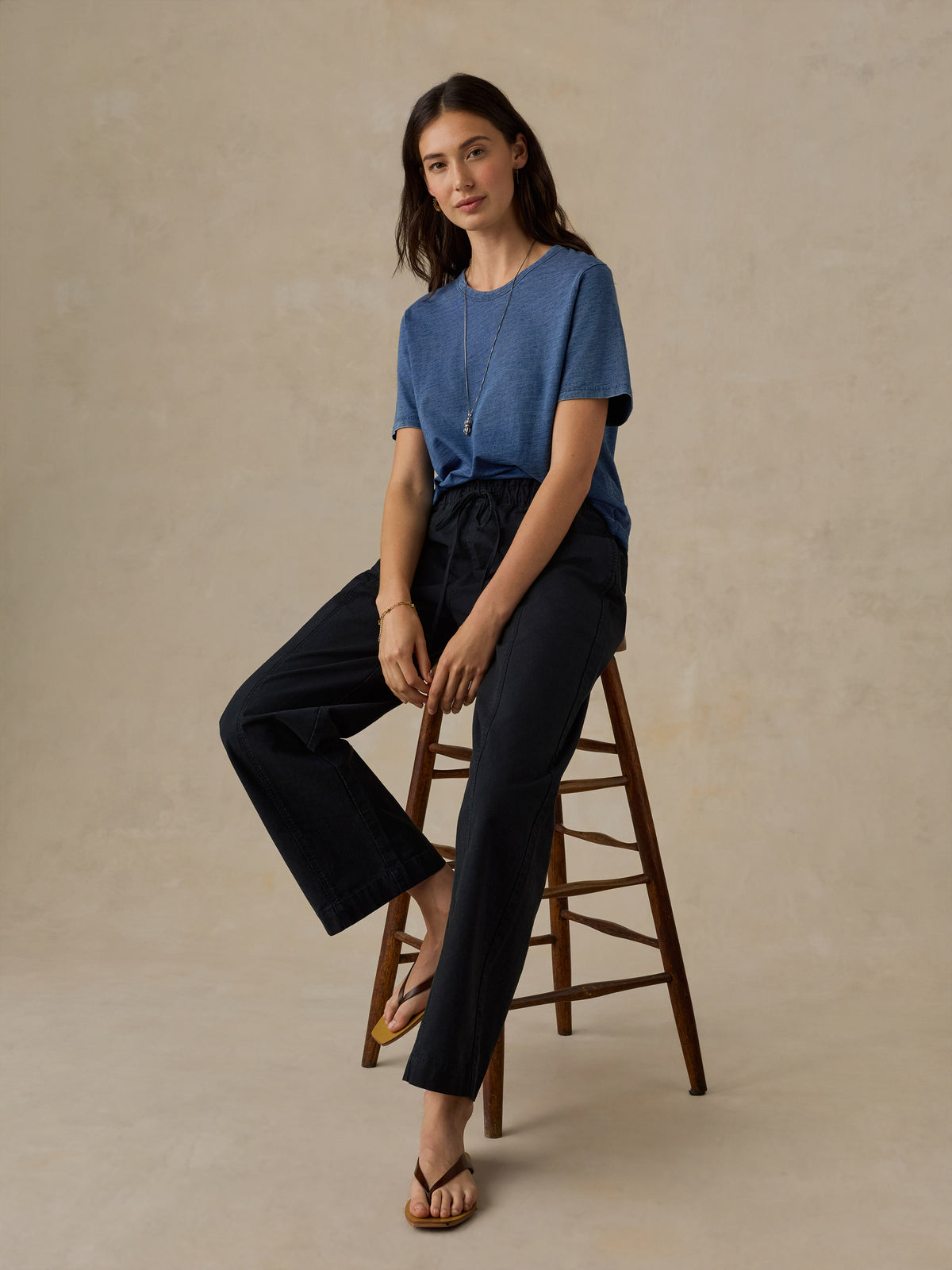 Woman sitting on a wooden stool wearing a blue t-shirt and black drawstring pants against a beige background, image 3 of 5