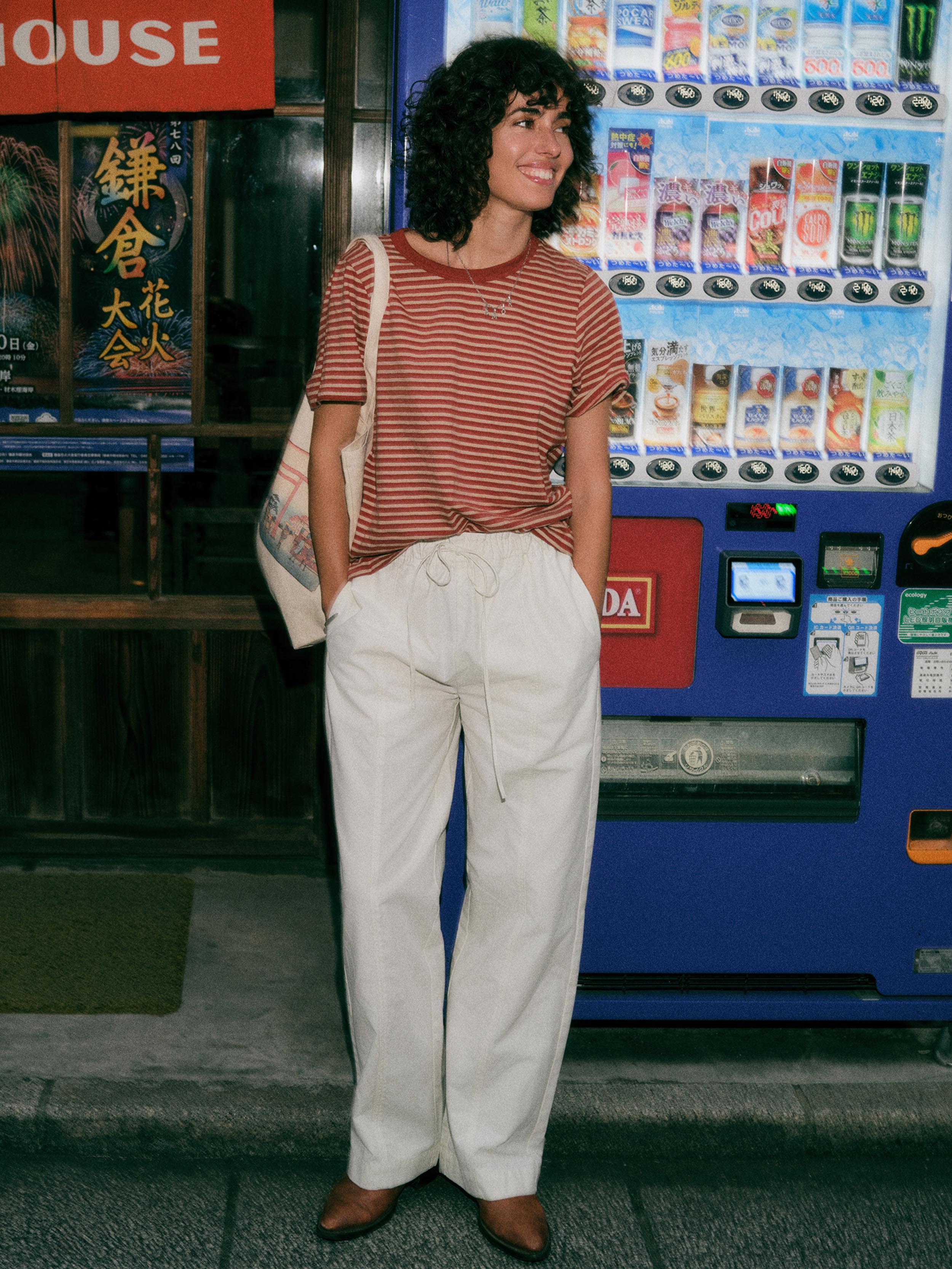 A woman stands in front of a vending machine wearing a rust-red and white striped t-shirt, cream wide-leg drawstring pants, and brown shoes, with a canvas tote bag slung over her shoulder.
