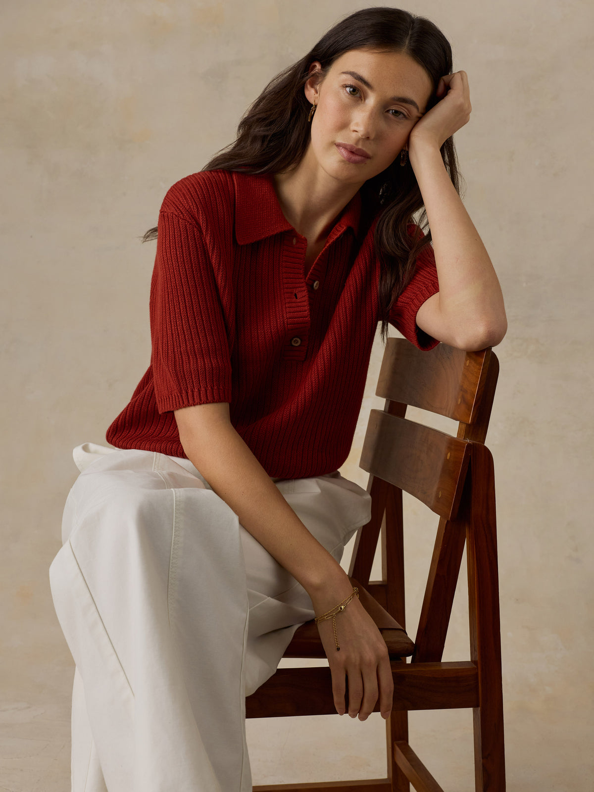 Woman sitting on a wooden chair wearing a red knit polo shirt and white drawstring pants against a beige background, image 4 of 6