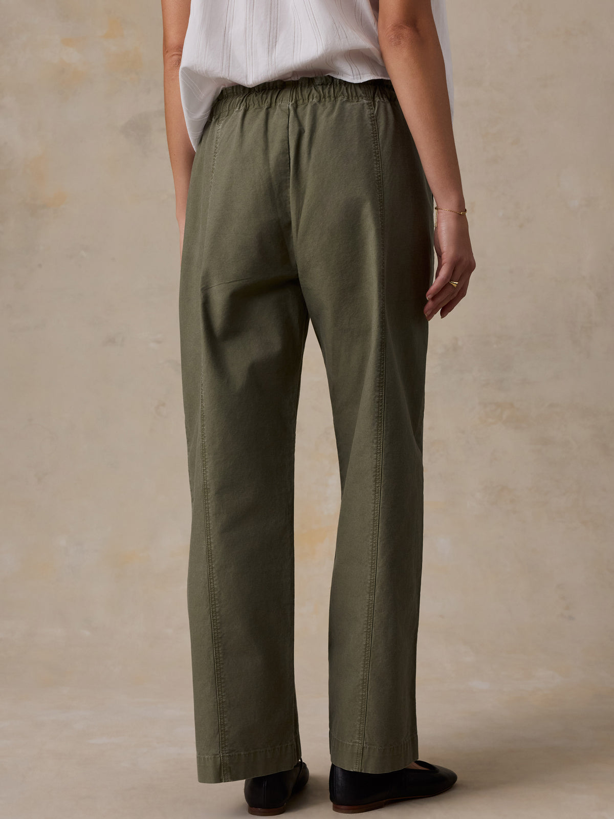 A woman shown from the back wearing a white blouse and olive green pants against a beige studio background, image 4 of 5