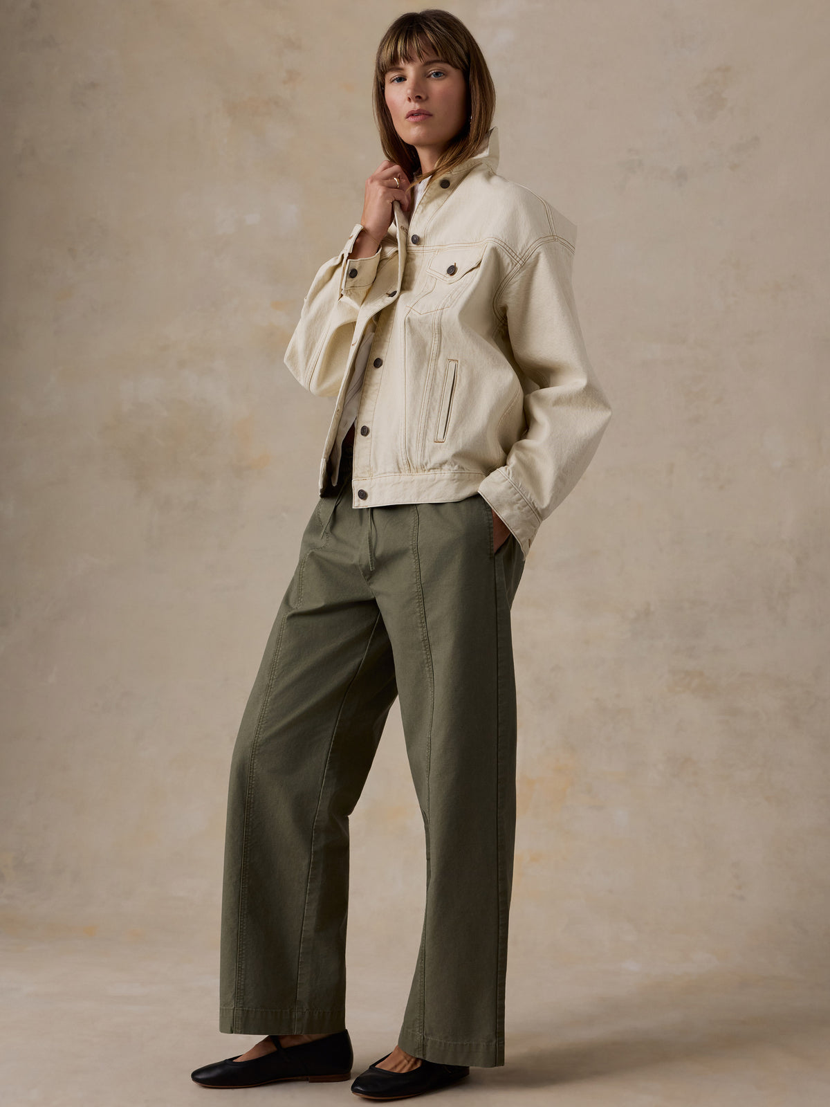 A woman wearing an off-white denim jacket and olive green pants against a beige studio background, image 1 of 5