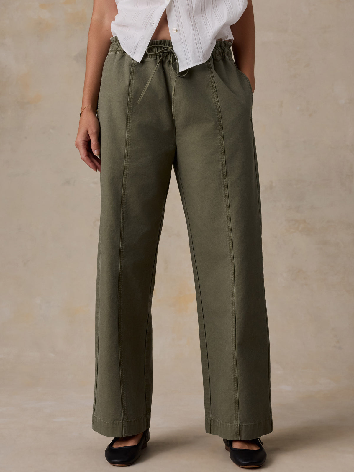 A woman wearing a white blouse and olive green pants against a beige studio background, image 2 of 5