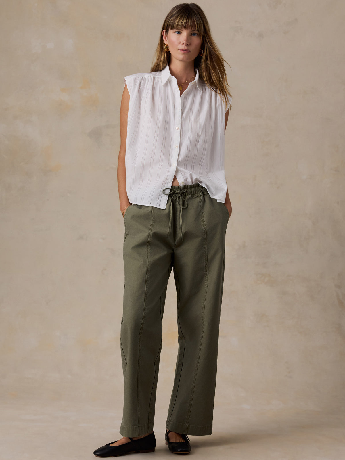 A woman wearing a white blouse and olive green pants against a beige studio background, image 3 of 5