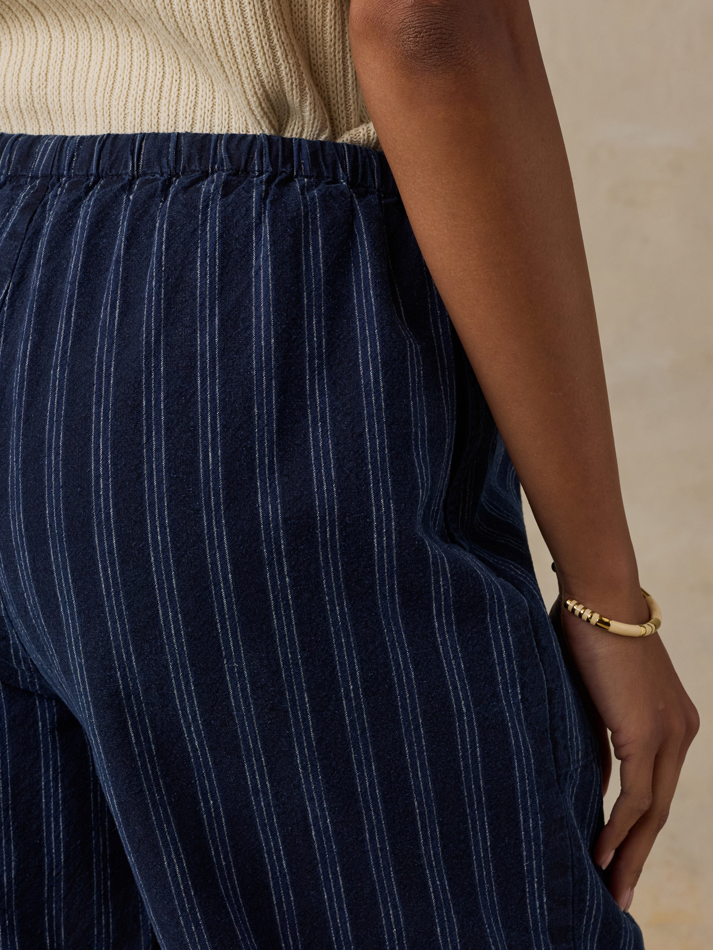 Close-up of a person wearing navy blue pinstriped trousers with a cream ribbed top and a gold bracelet, set against a neutral beige background.