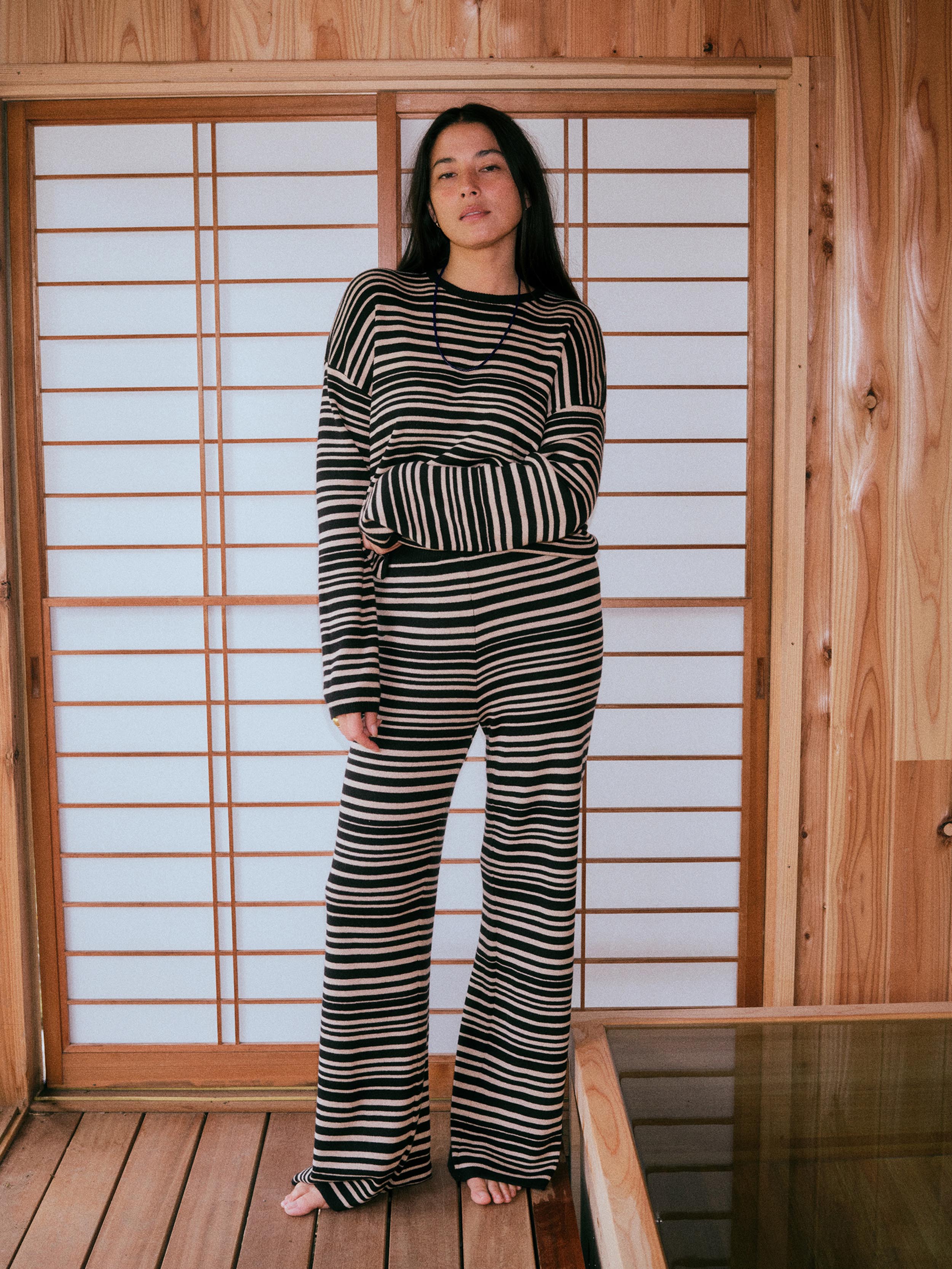 A woman stands barefoot in front of a wooden shoji screen, wearing a matching black and beige striped long-sleeve top and wide-leg pants set.