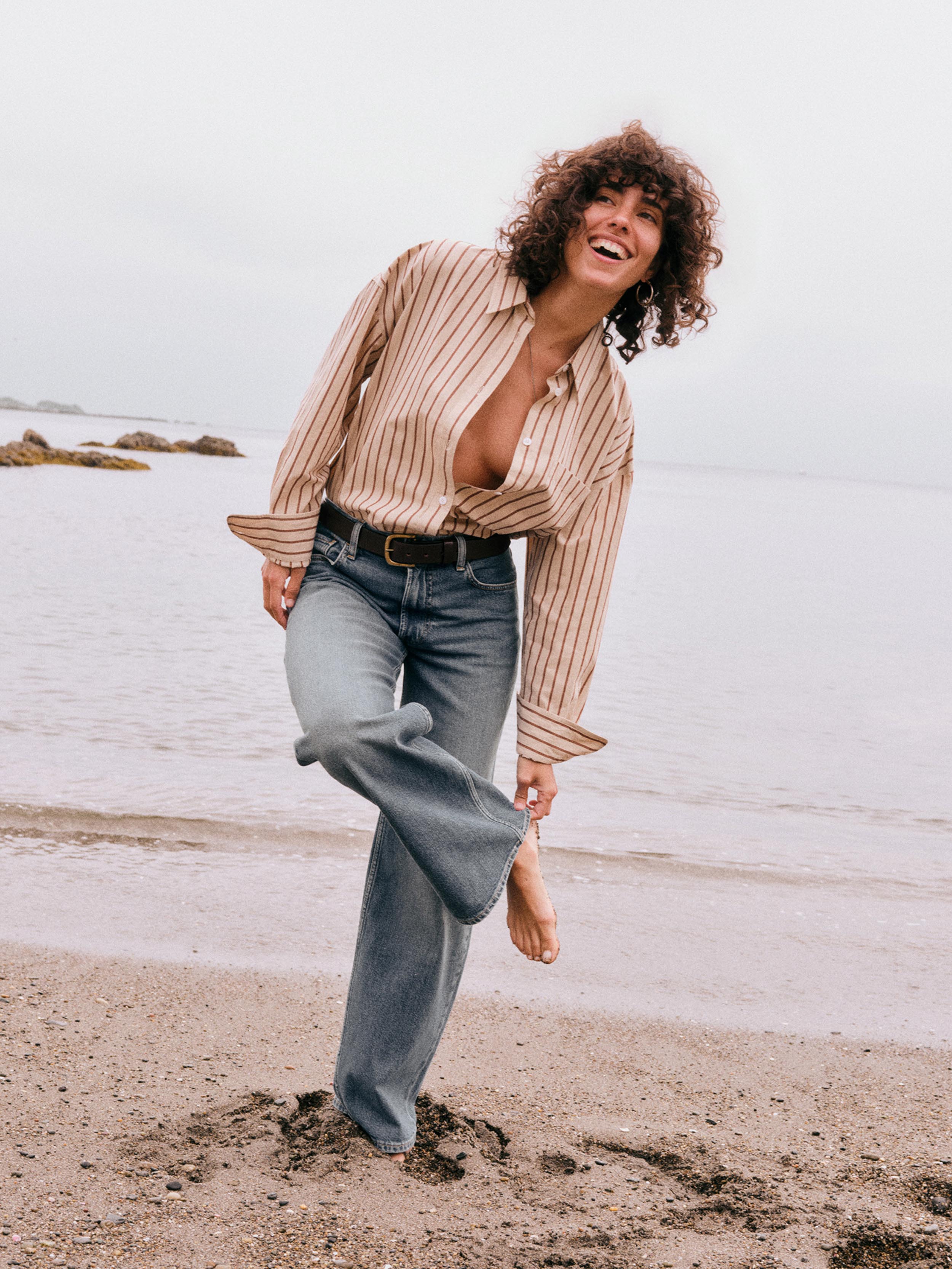A woman with curly brown hair stands barefoot on a sandy beach, wearing a beige and brown vertically striped button-down shirt, blue wide-leg jeans, and a black belt. She is smiling and looking up towards the overcast sky.