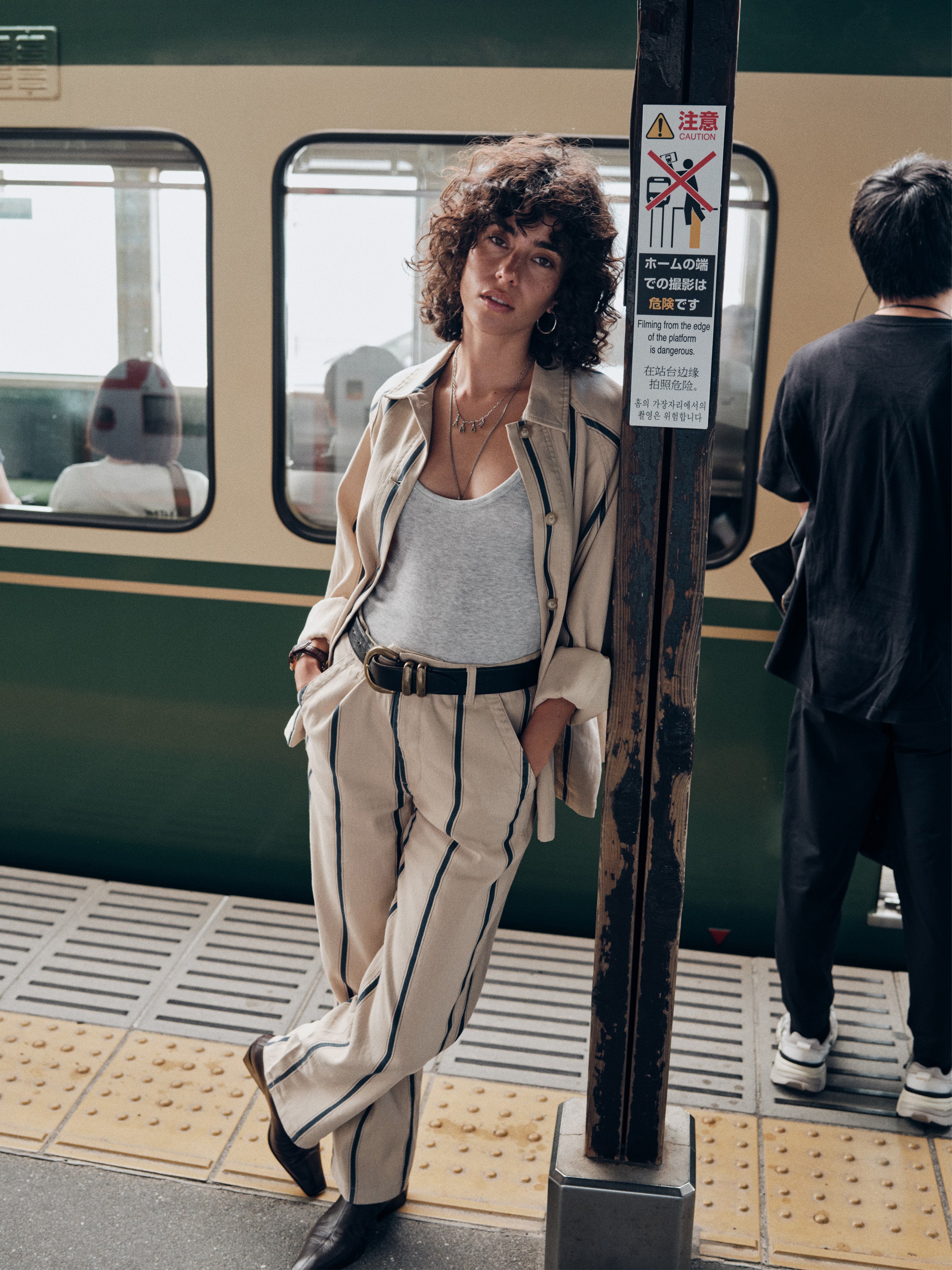A woman stands on a subway platform wearing a beige striped suit with a white tank top and black belt, posing next to a train and a metal pole.
