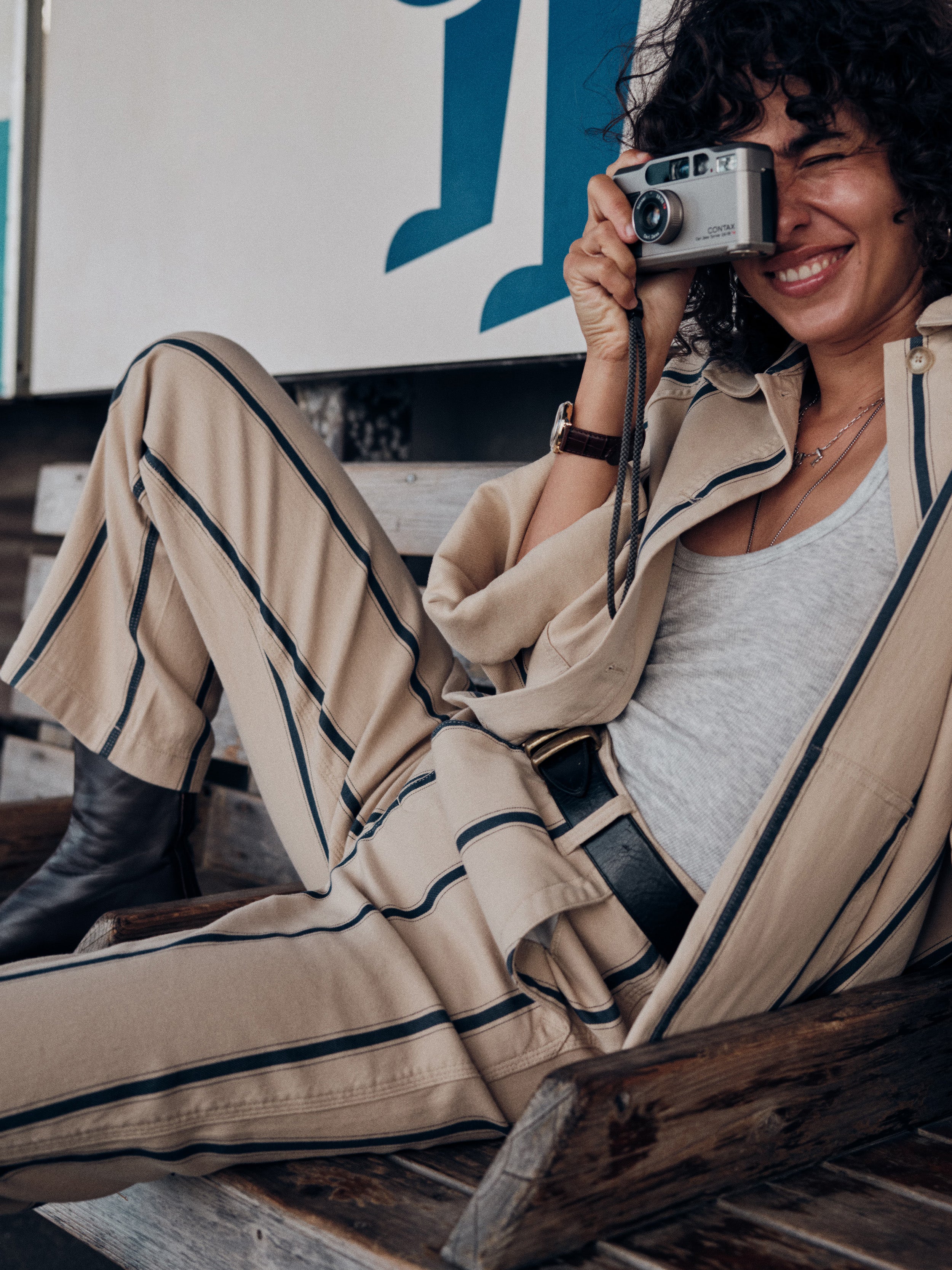 A woman sitting on a wooden bench wearing a beige matching set with black stripes, holding a silver camera to her eye.