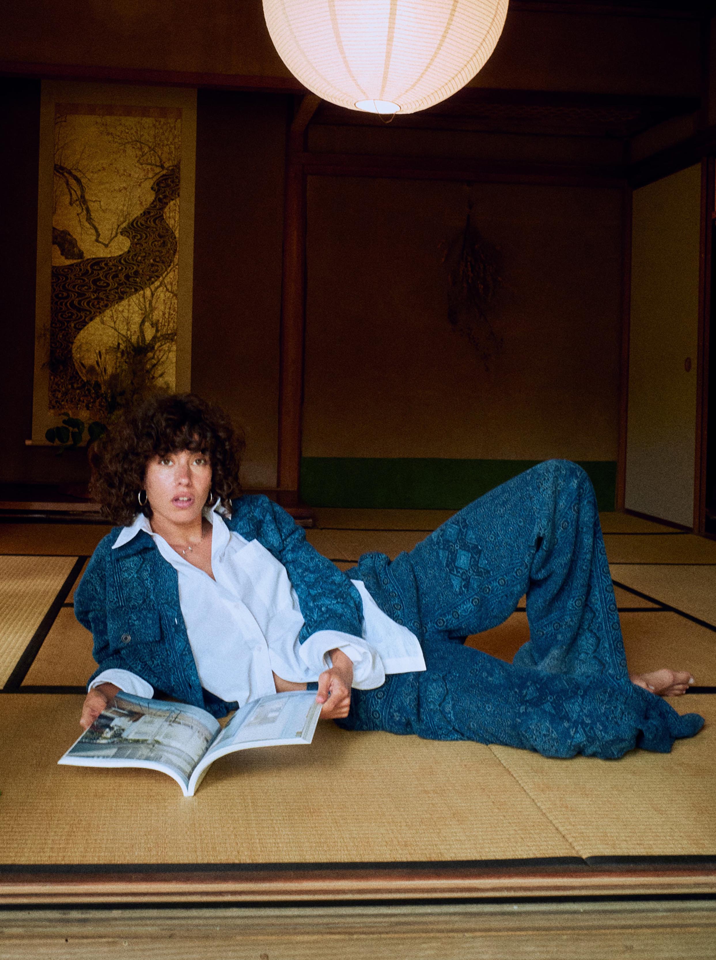 A woman reclining on a tatami floor in a traditional Japanese room, wearing a blue patterned jacket and matching wide-leg pants over a white shirt, while holding an open magazine.
