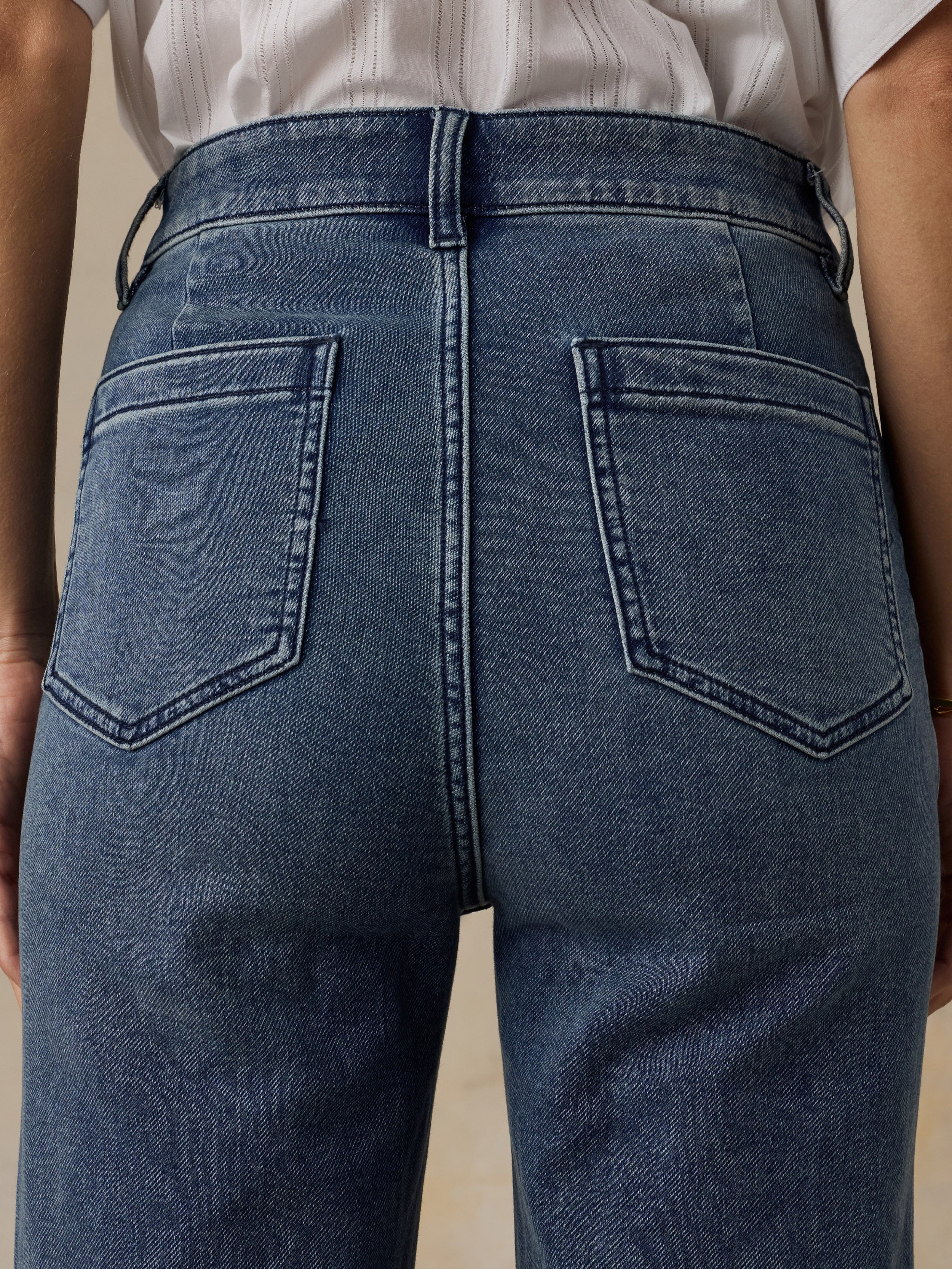 Close-up of a woman wearing a white top and high waisted blue jeans against a beige background