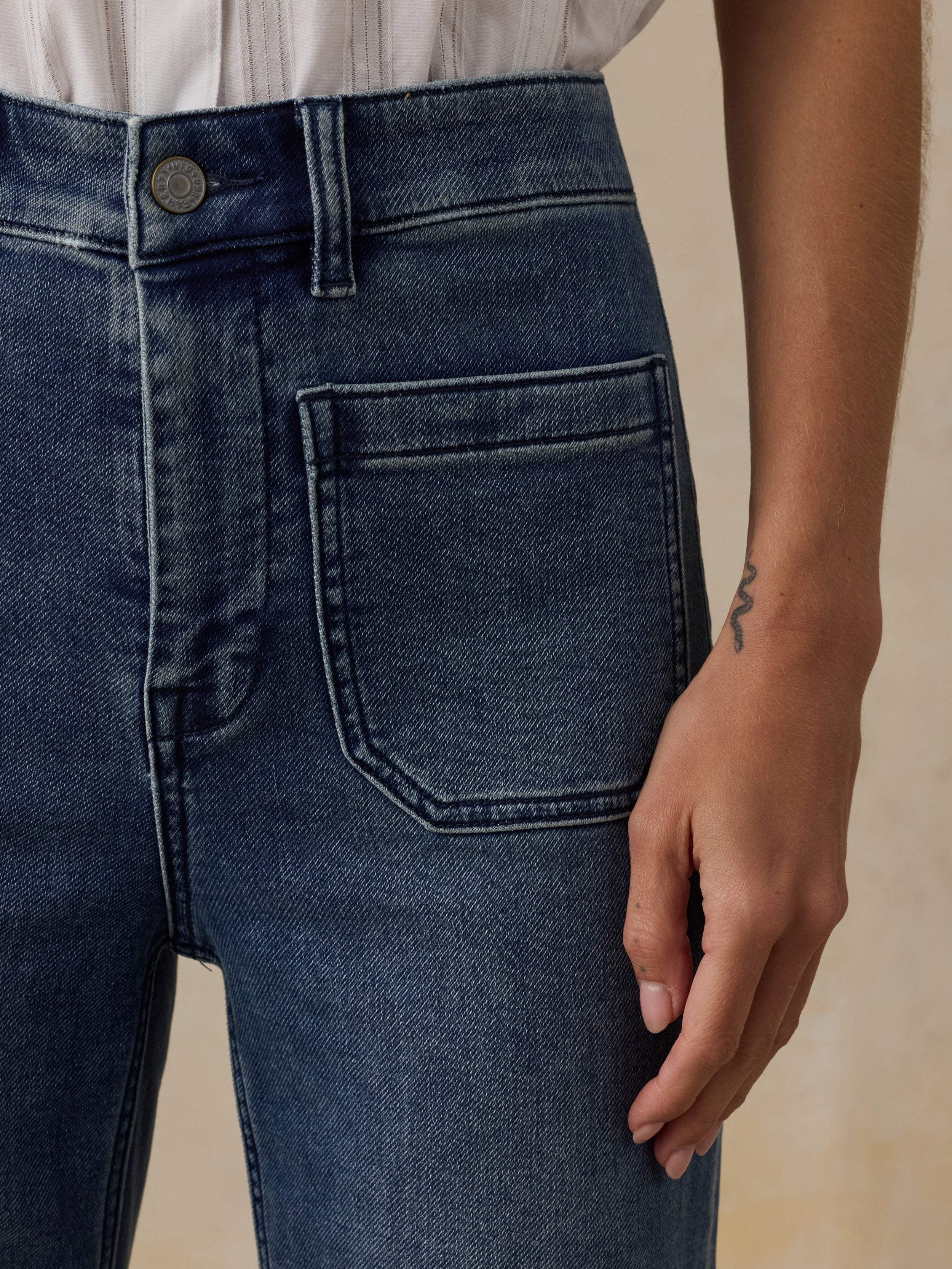 Close-up of a woman wearing a white top and high waisted blue jeans against a beige background