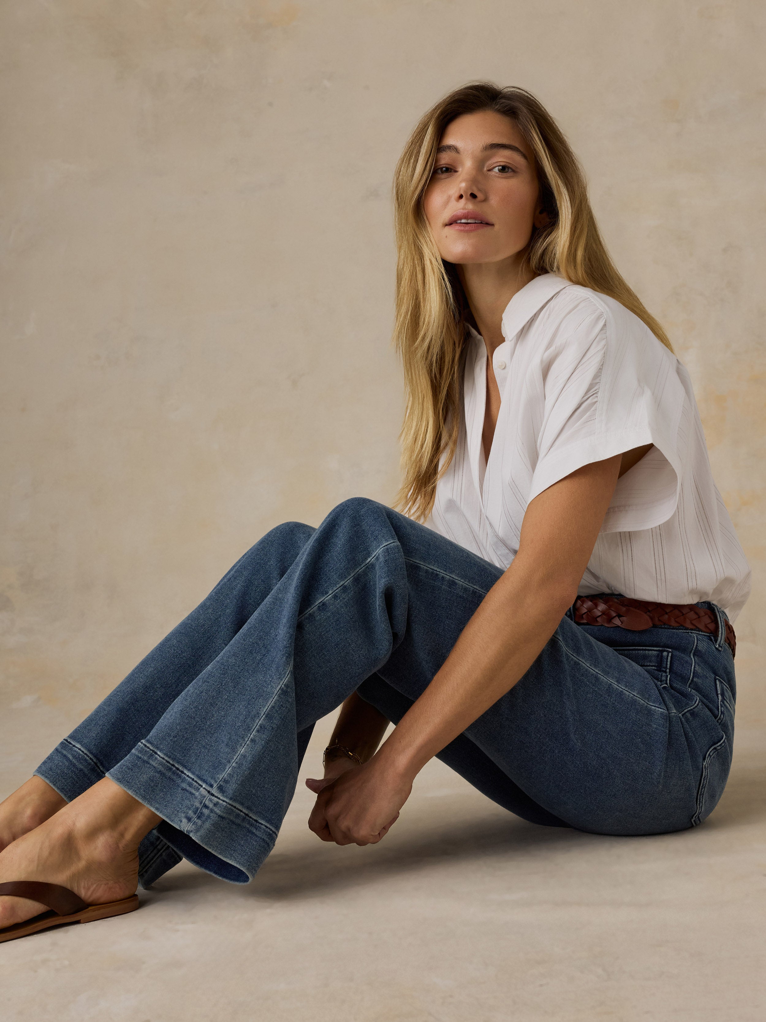 Woman sitting on the floor wearing a white top and high waisted blue jeans against a beige background