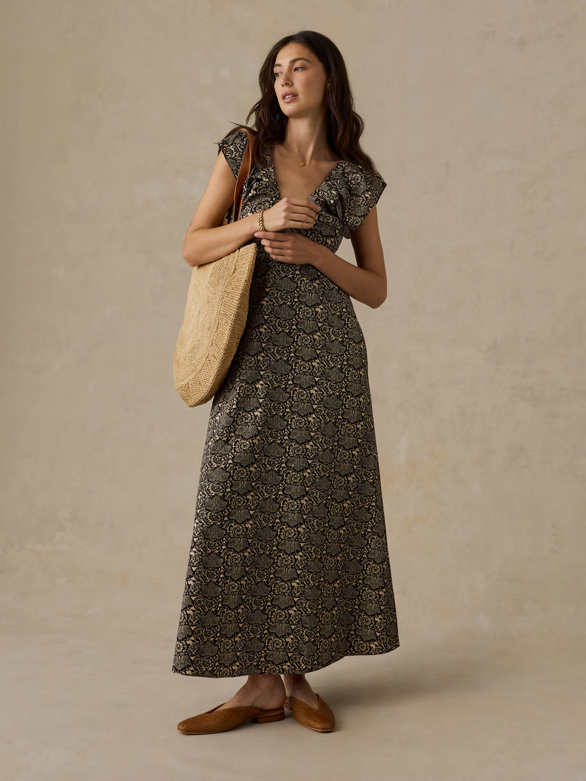 Woman wearing a black and beige floral printed dress and raffia bag against a beige background, image 2 of 6