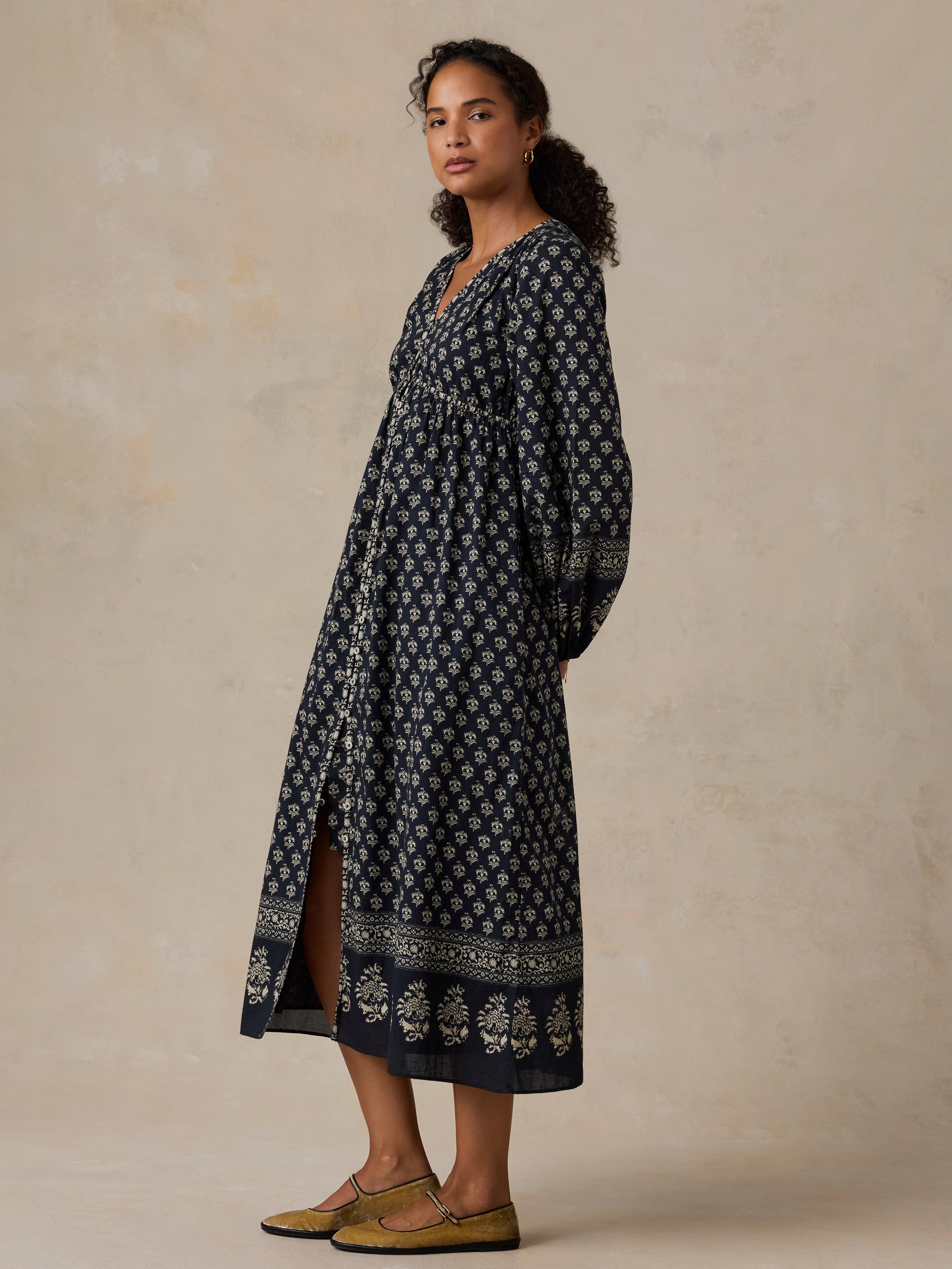 A woman modeling a long-sleeved navy blue maxi dress with a white floral block print and a decorative border along the hem, paired with olive green flat shoes against a textured beige studio backdrop.
