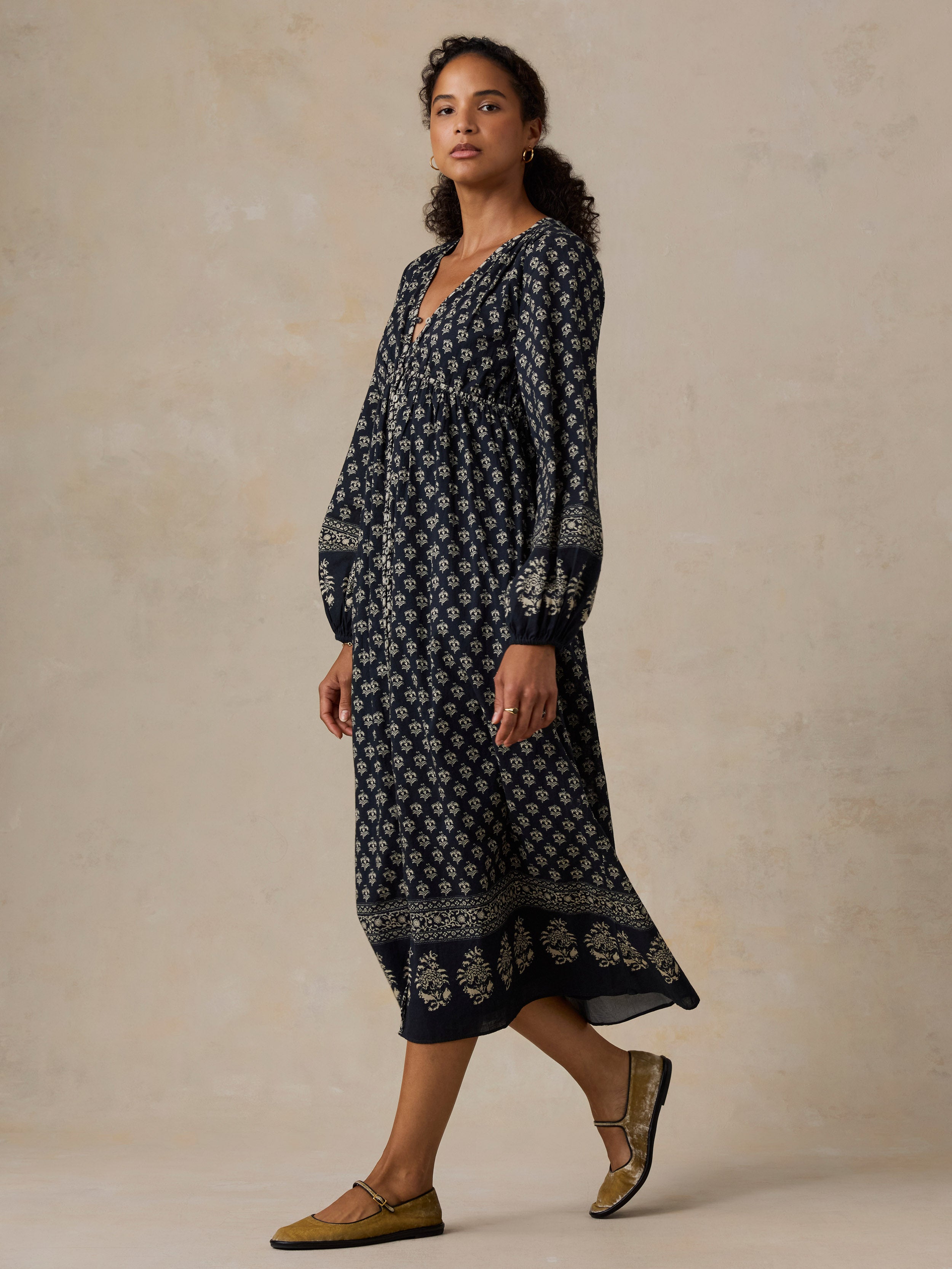 A woman modeling a long-sleeve navy midi dress with a white floral block print and a decorative border, paired with olive green flat sandals, set against a textured beige studio backdrop.