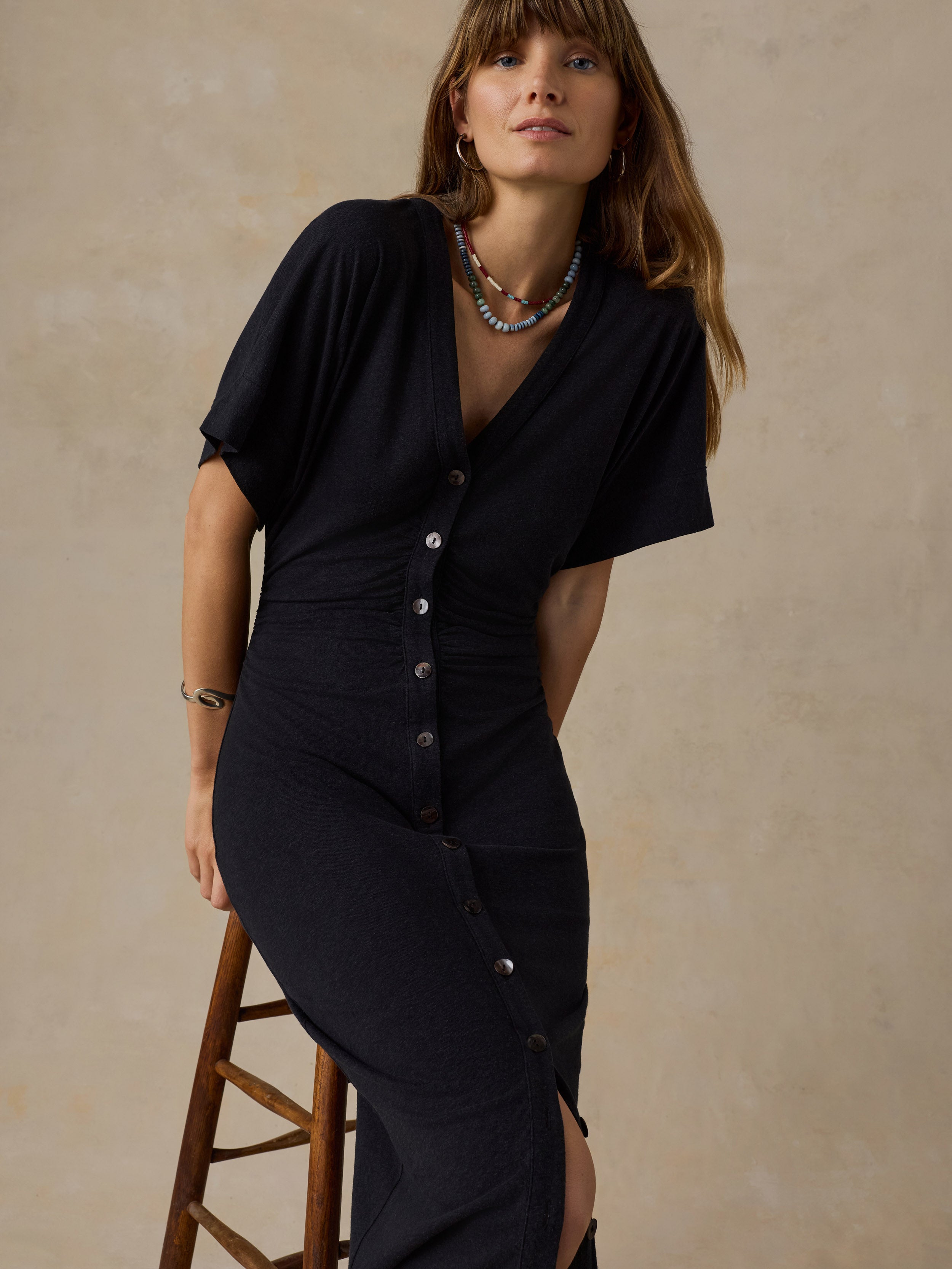 Woman sitting on a wooden stool wearing a black maxi dress against a beige background