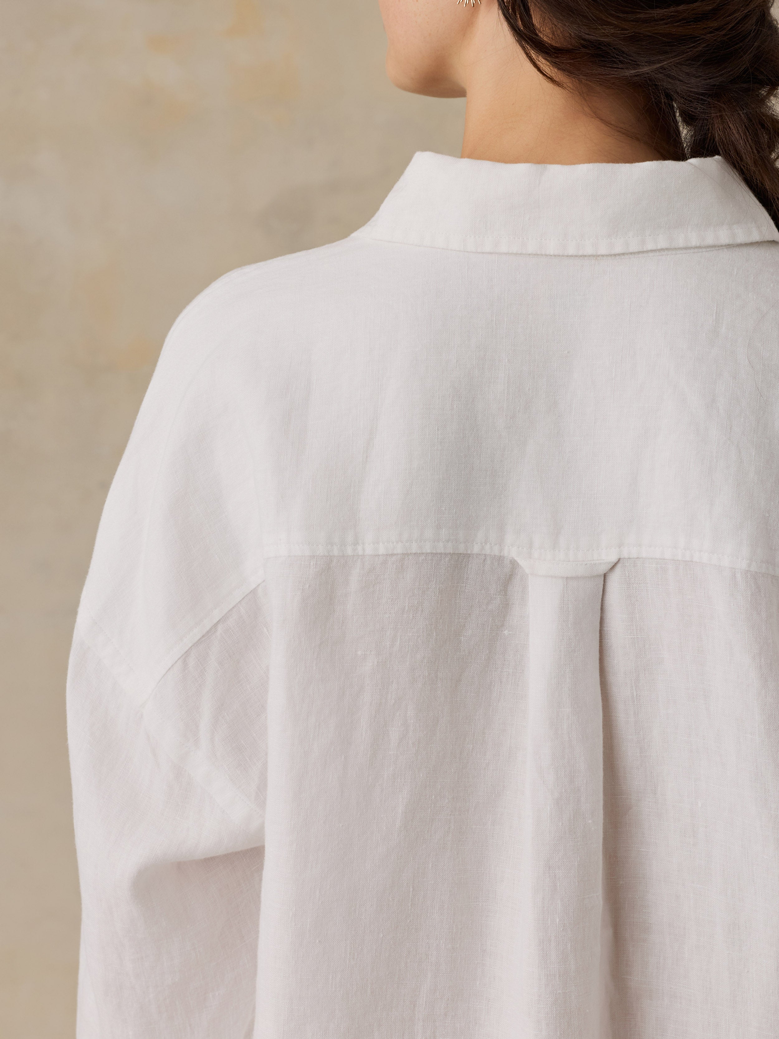Close-up back view of a woman wearing an oversized white linen shirt with a collar and gathered back seam, against a warm beige textured background.