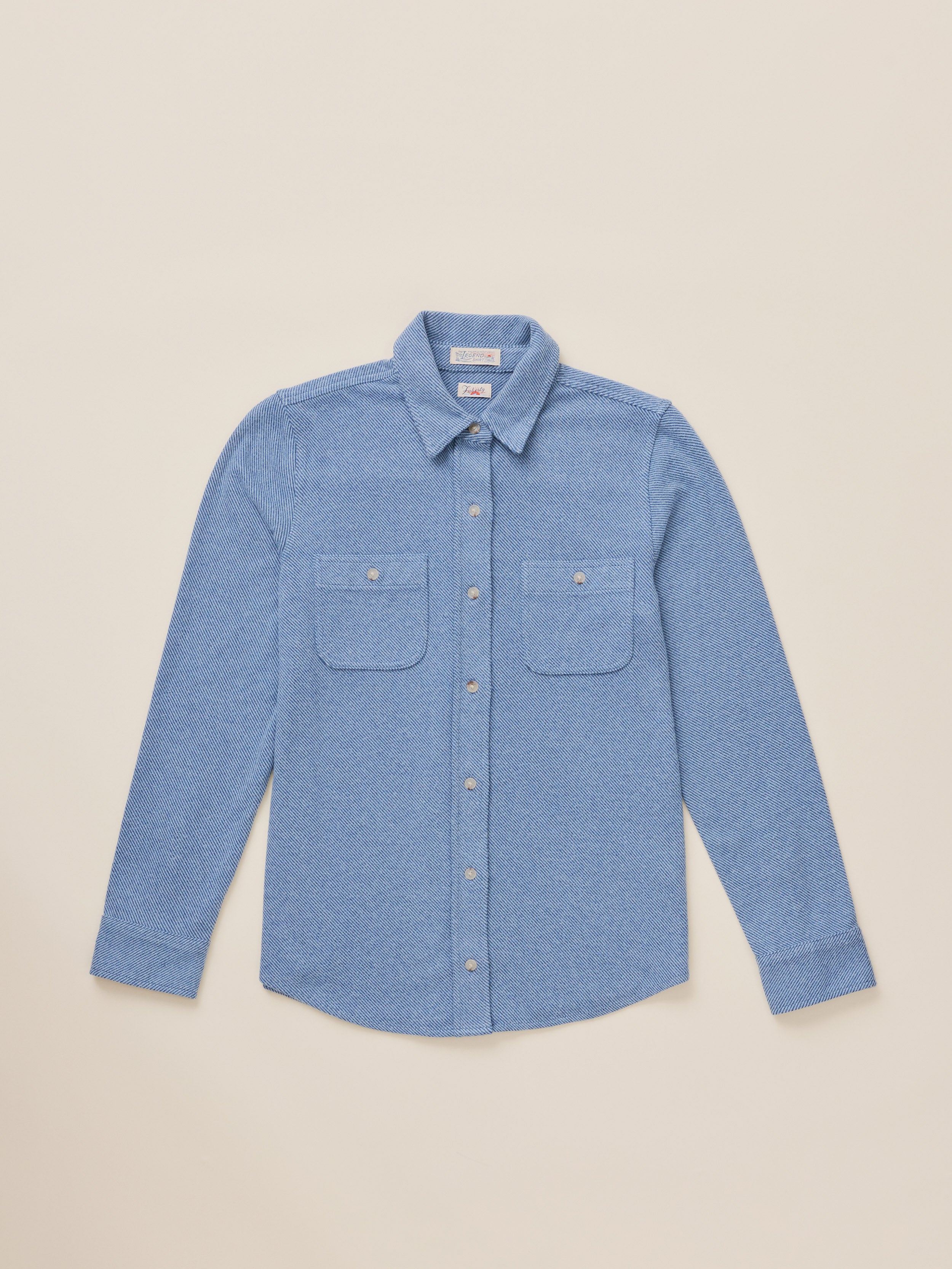 A light blue long-sleeve button-up shirt with two chest pockets, laid flat against a solid beige background.