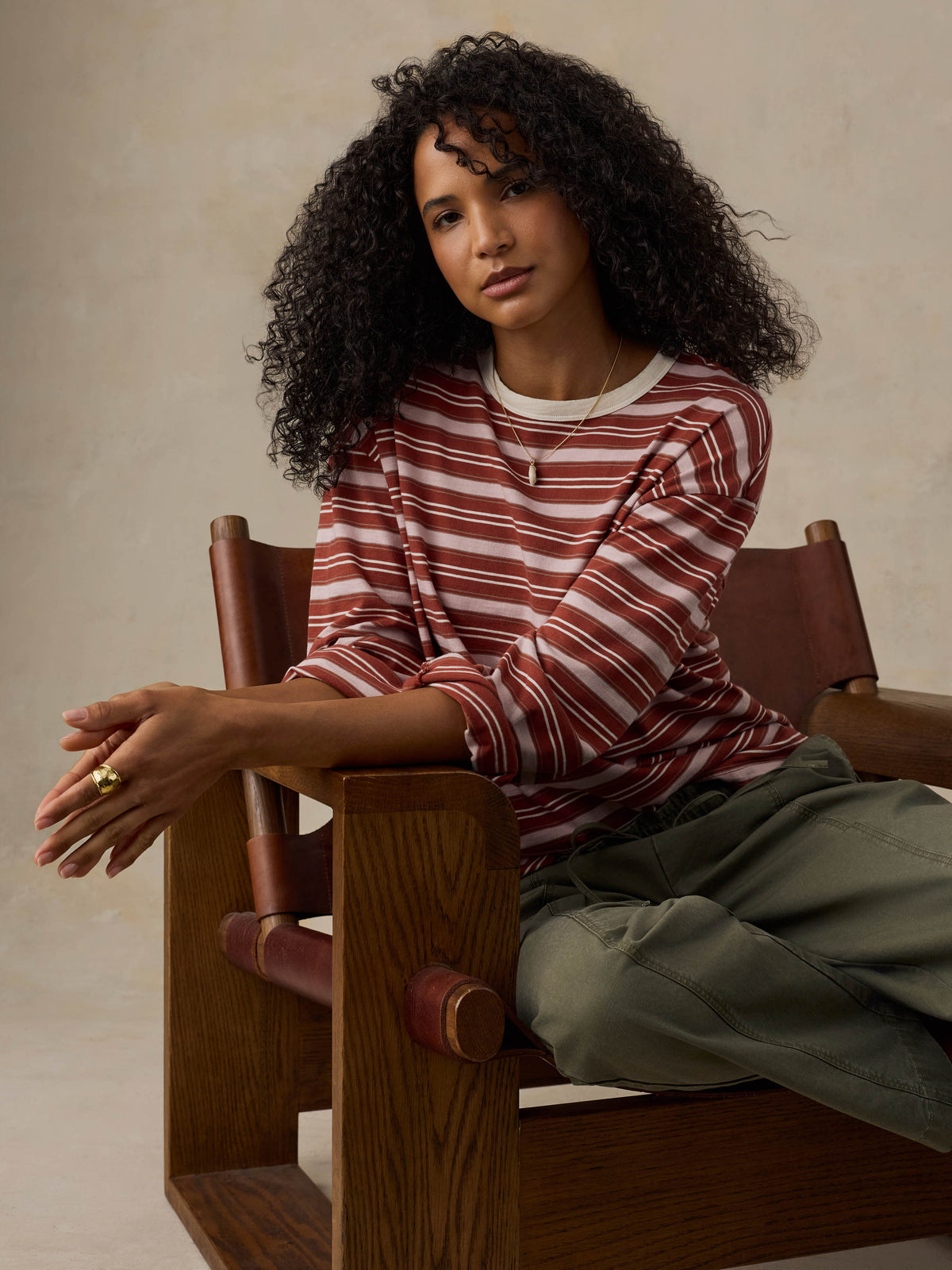 Woman sitting on a wooden chair wearing a red and pink striped long-sleeve shirt and green pants against a beige background, image 4 of 7