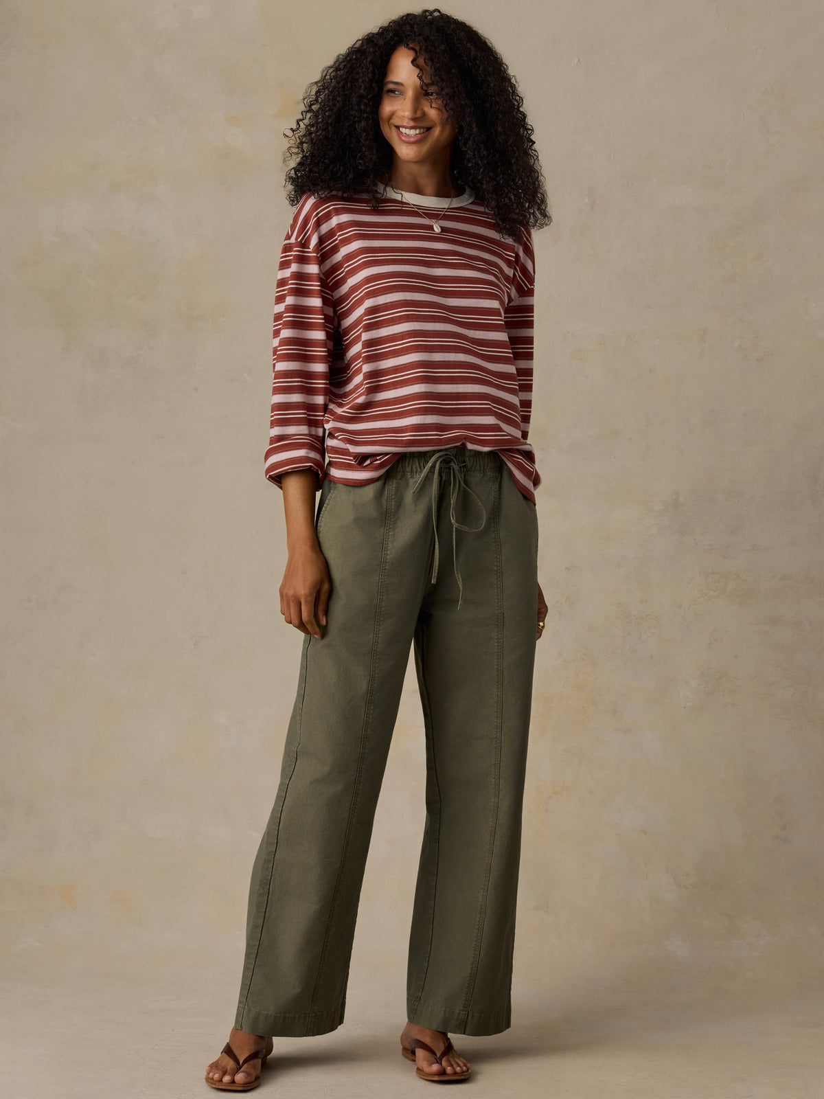 Woman wearing a red and pink striped long-sleeve shirt and green pants against a beige background, image 6 of 7