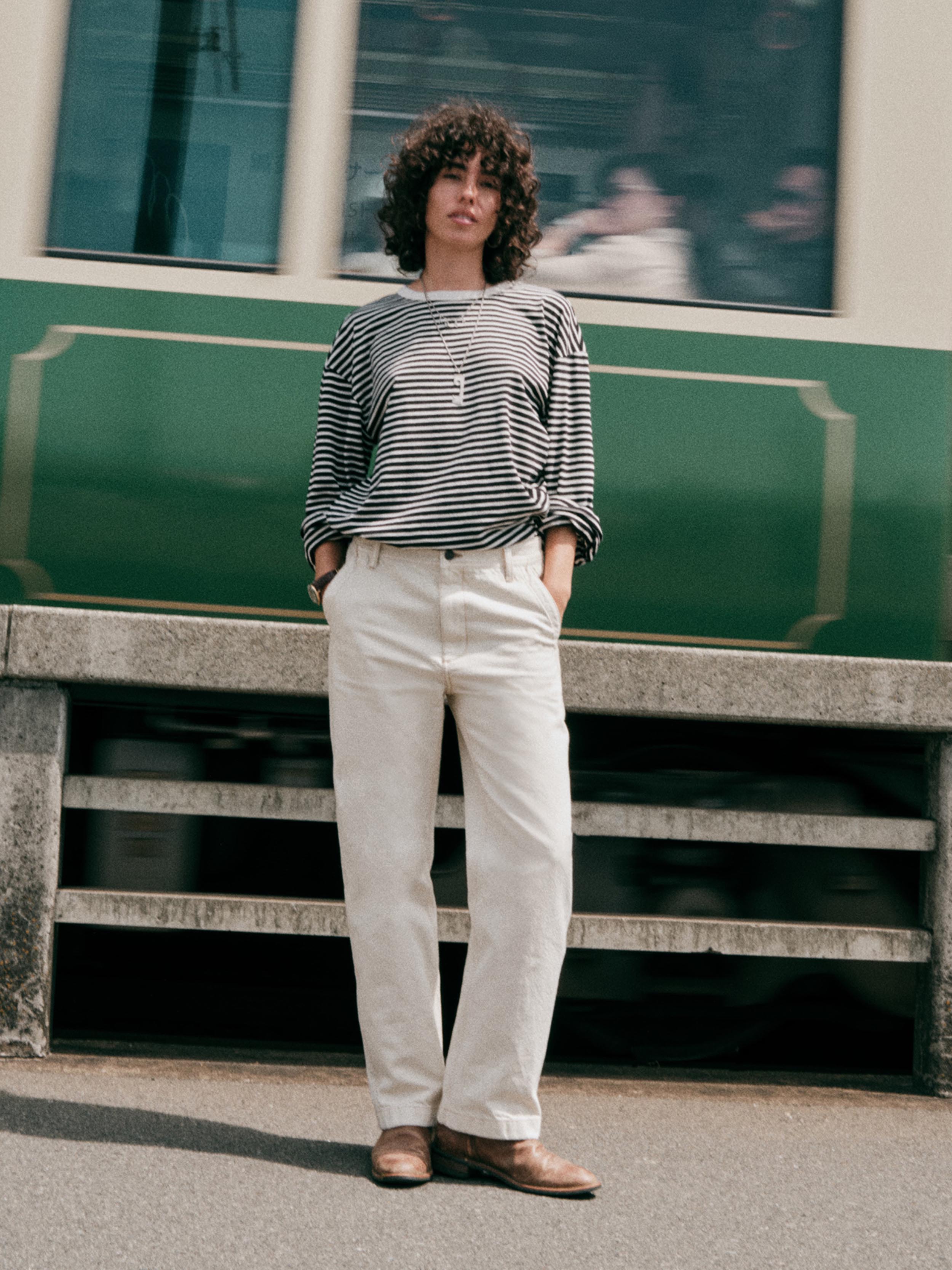 A woman stands outdoors in front of a green and cream train, wearing a black and white striped long-sleeve top, off-white trousers, and brown leather shoes.