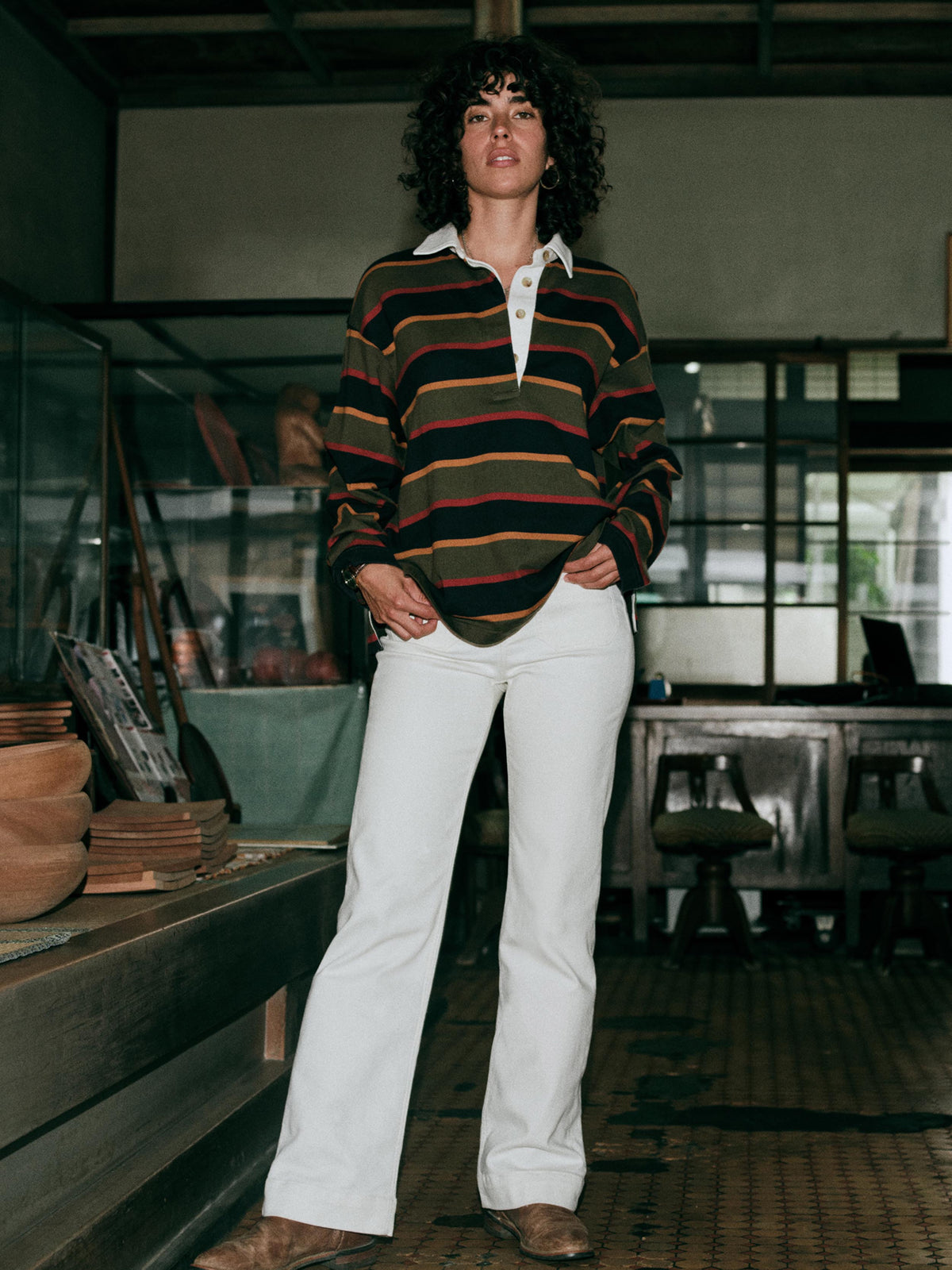 A woman wearing a long-sleeve rugby-style shirt with stripes in olive green, navy blue, red, and mustard yellow and white pants, image 7 of 7