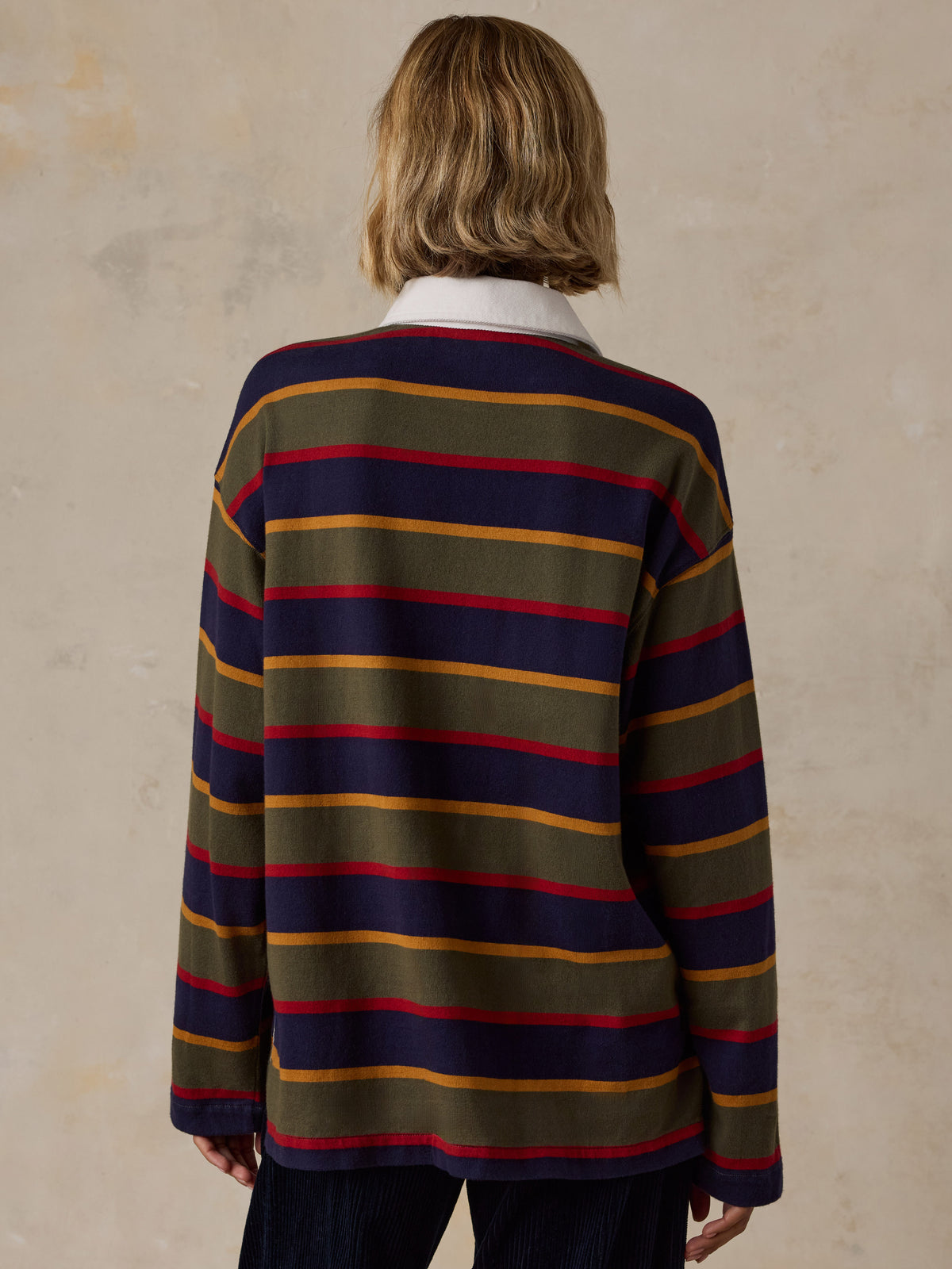 A woman wearing a long-sleeve rugby-style shirt with stripes in olive green, navy blue, red, and mustard yellow, image 4 of 7