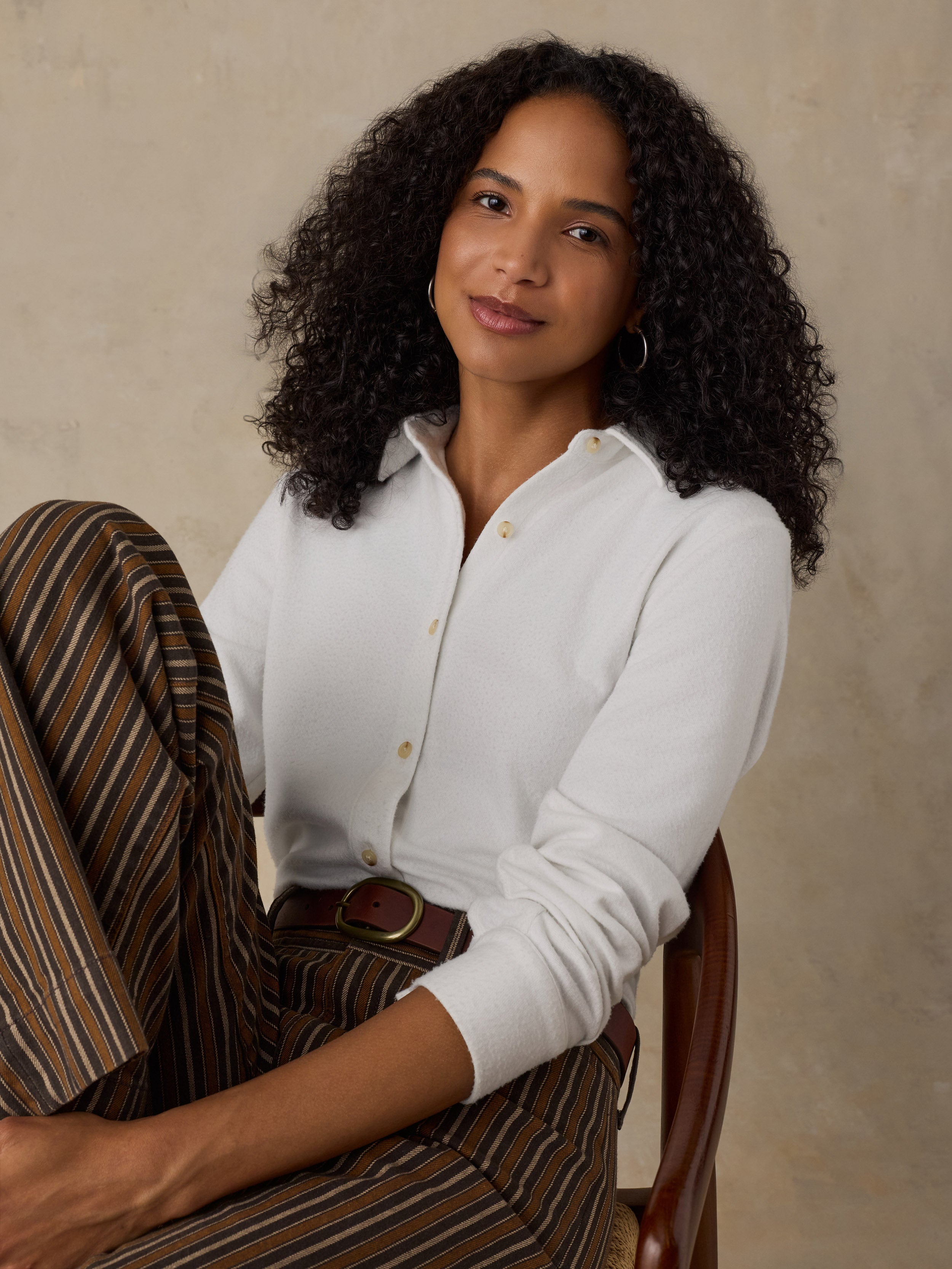 Woman sitting on a wooden chair wearing a white button up shirt and brown striped pants against a beige background
