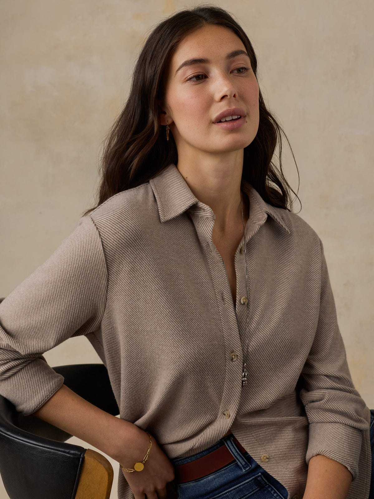 Woman sitting on a wooden chair wearing a beige button down shirt and blue jeans against a beige background, image 2 of 6