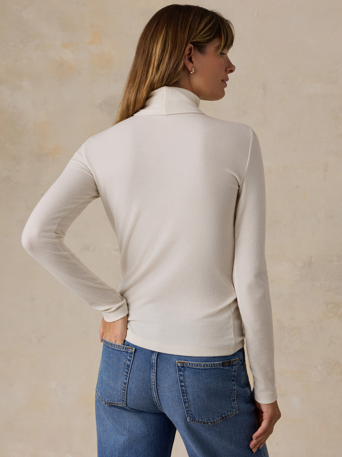 A woman viewed from behind wearing a white turtleneck and blue jeans, image 4 of 7