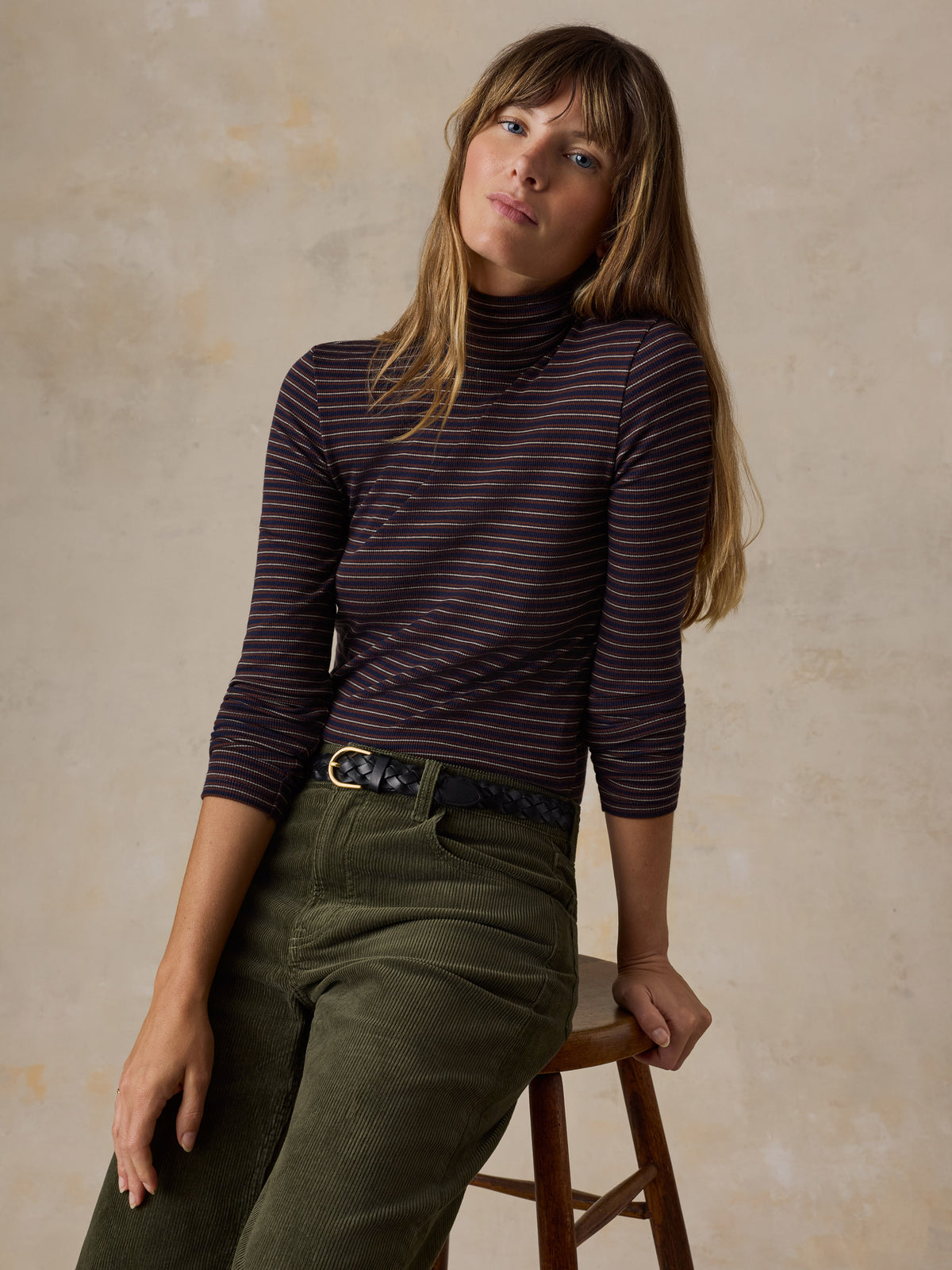 A woman sitting on a stool wearing a brown striped turtleneck and green corduroy pants, image 4 of 6