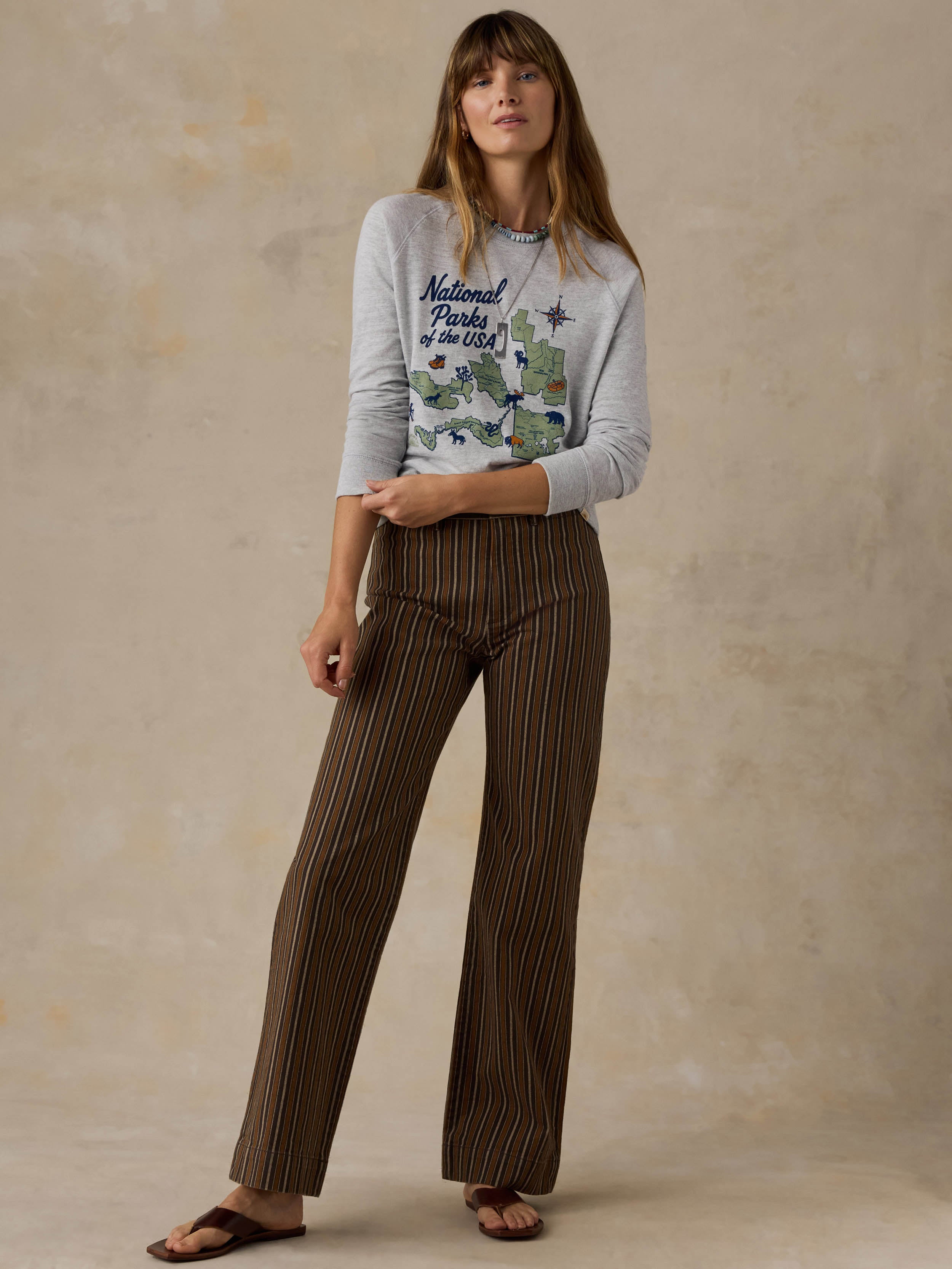 Woman wearing a grey crewneck with a National Parks graphic on the front and brown striped pants against a beige background