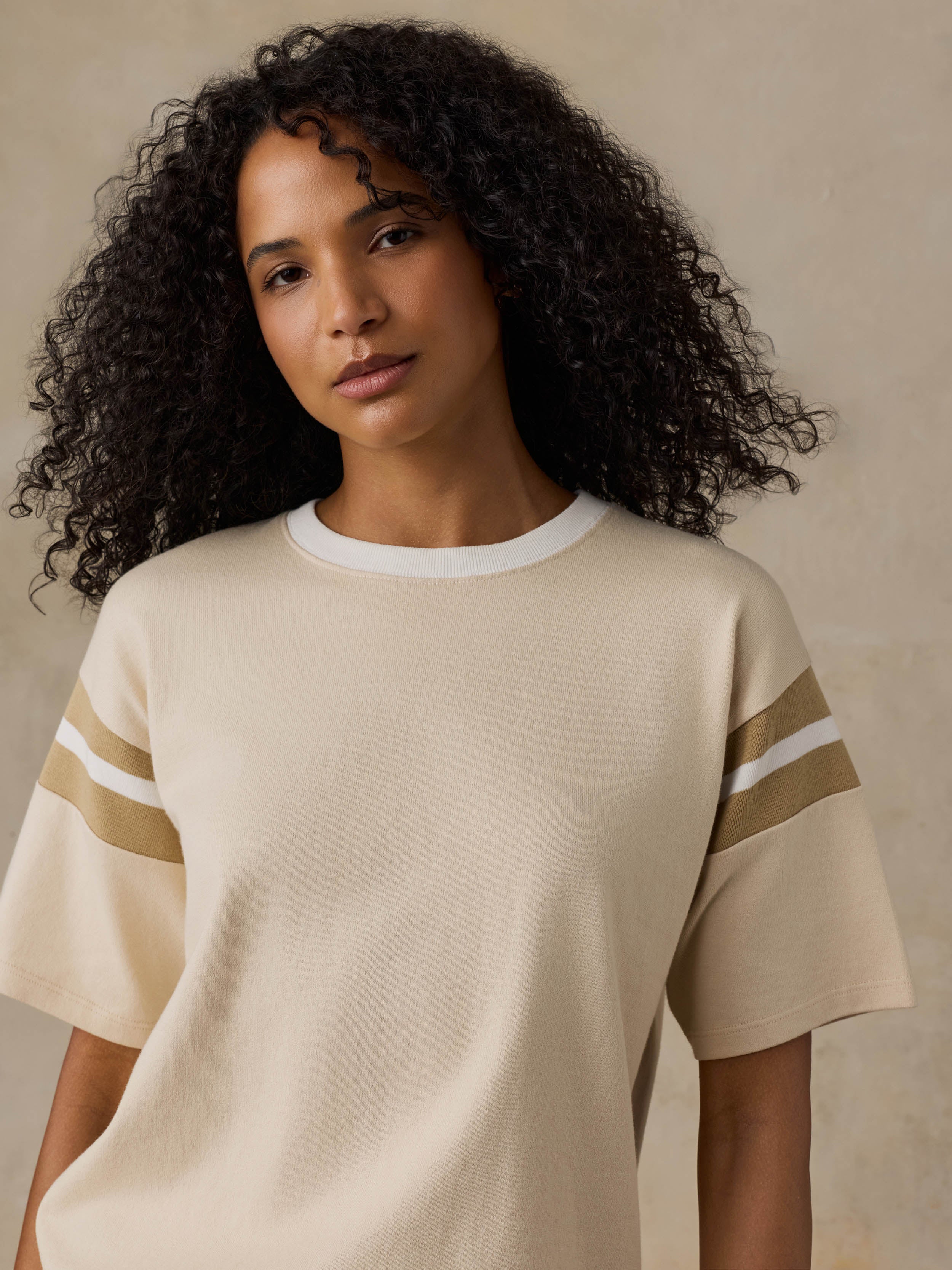 A woman with curly black hair wearing a cream-colored short-sleeve crewneck top with white and tan stripes on the sleeves, set against a neutral beige background.