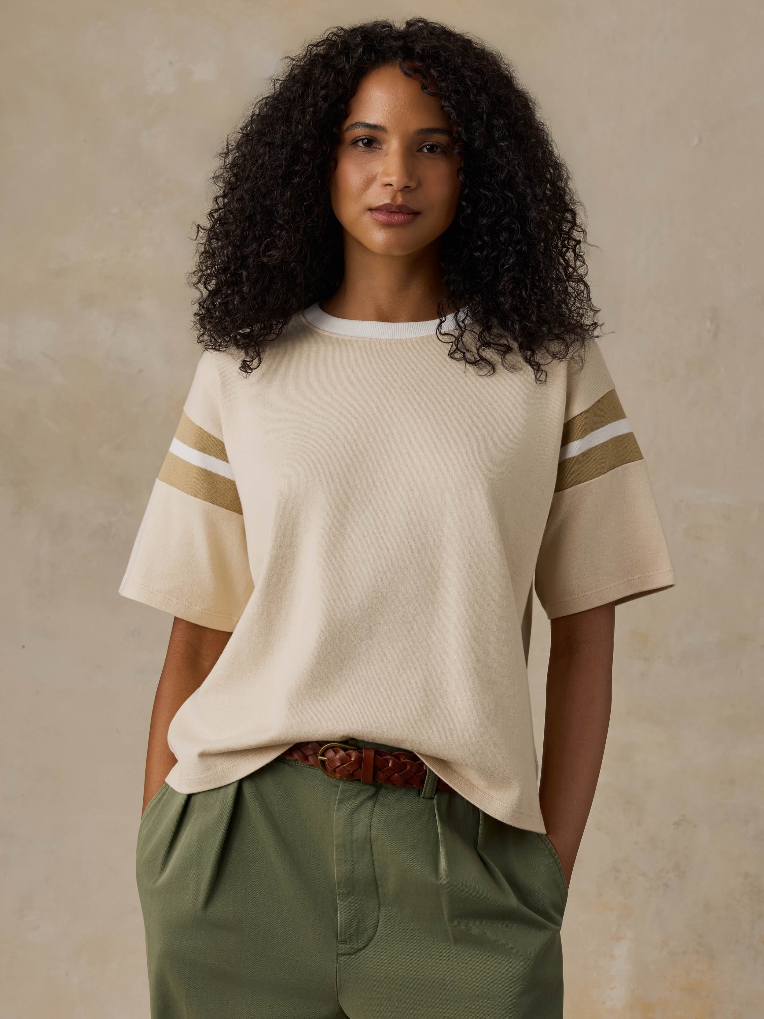 A woman with curly black hair wearing a cream short-sleeve knit top with beige stripes on the sleeves, paired with olive green pants and a brown belt, standing against a textured beige background.