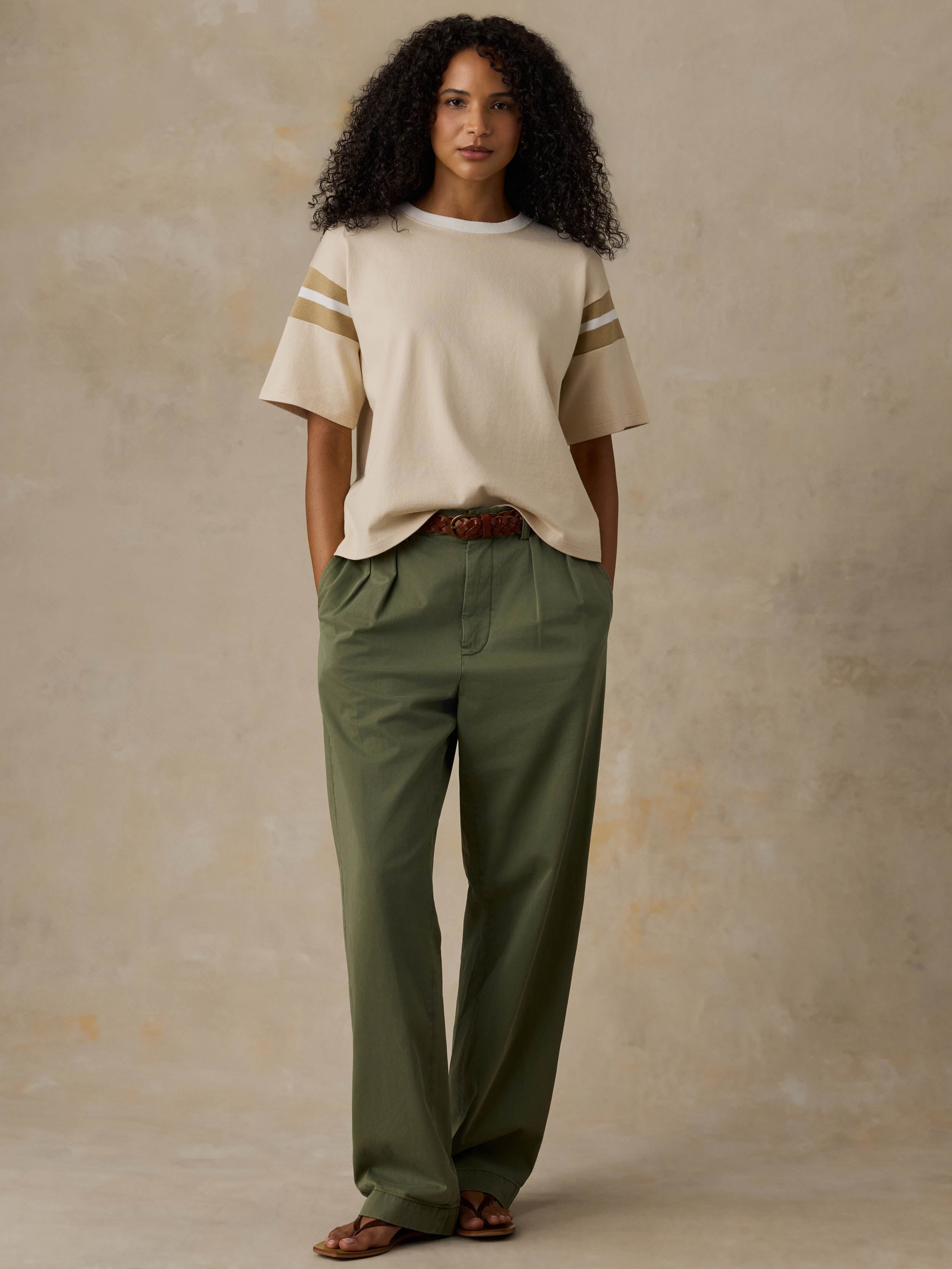 A woman stands against a textured beige studio backdrop wearing a cream short-sleeve top with beige stripes on the sleeves, olive green pants, and brown sandals.