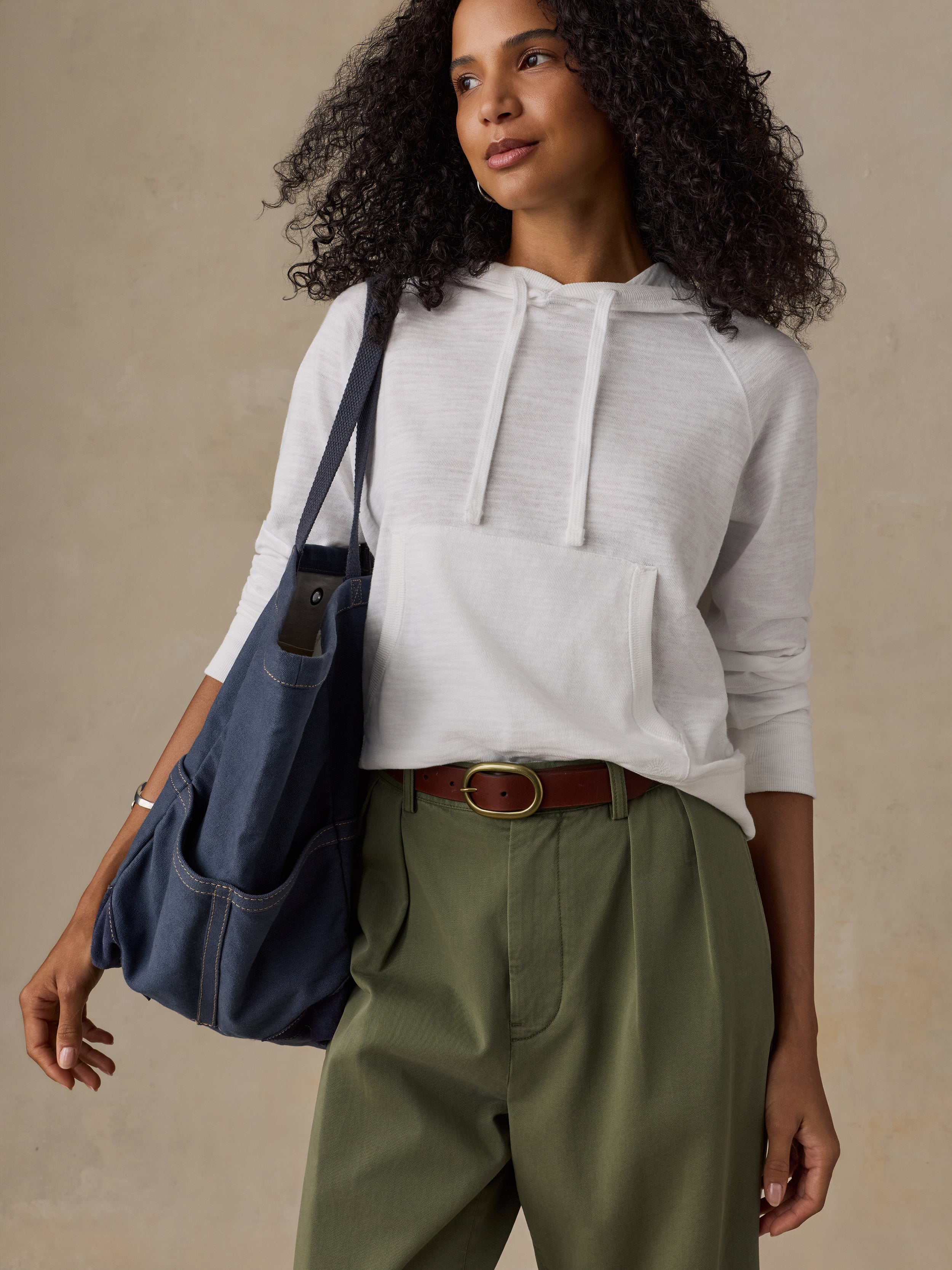 Woman wearing a white hoodie and olive green pants while carrying a denim tote bag against a beige background