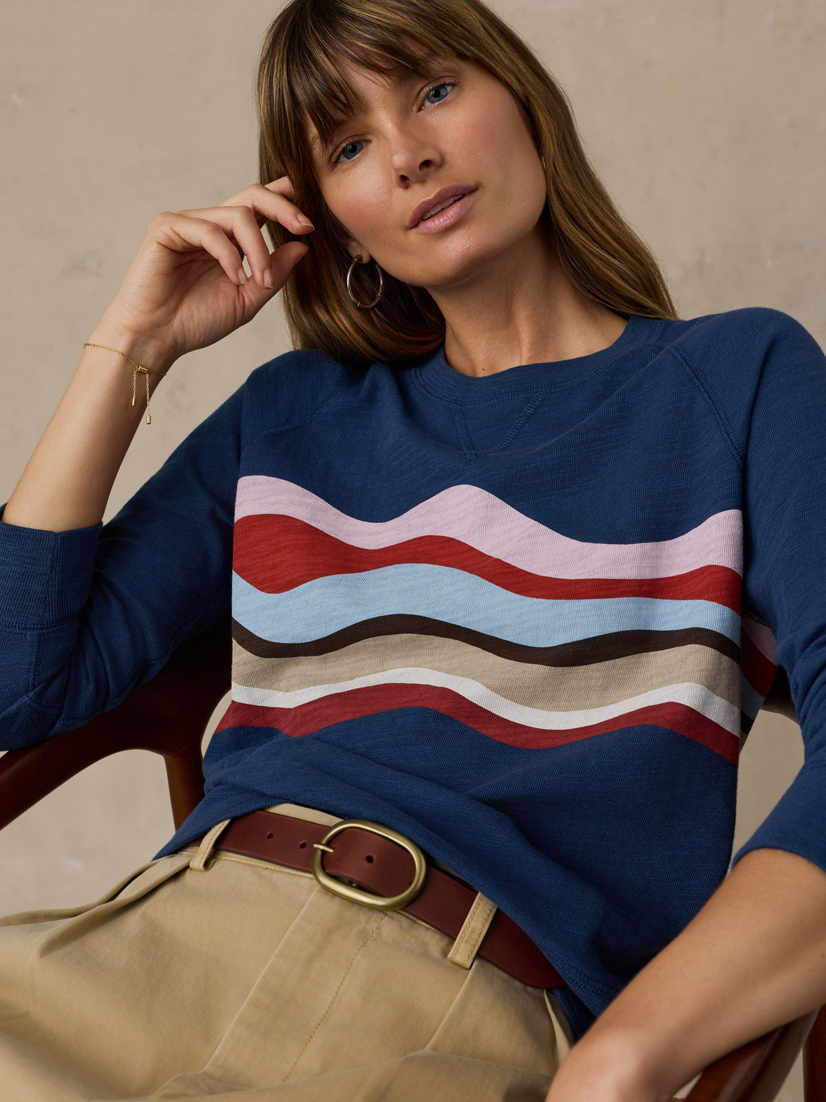 Woman sitting on a wooden chair wearing a navy blue and multicolor graphic crewneck with beige chino pants against a beige background, image 2 of 6