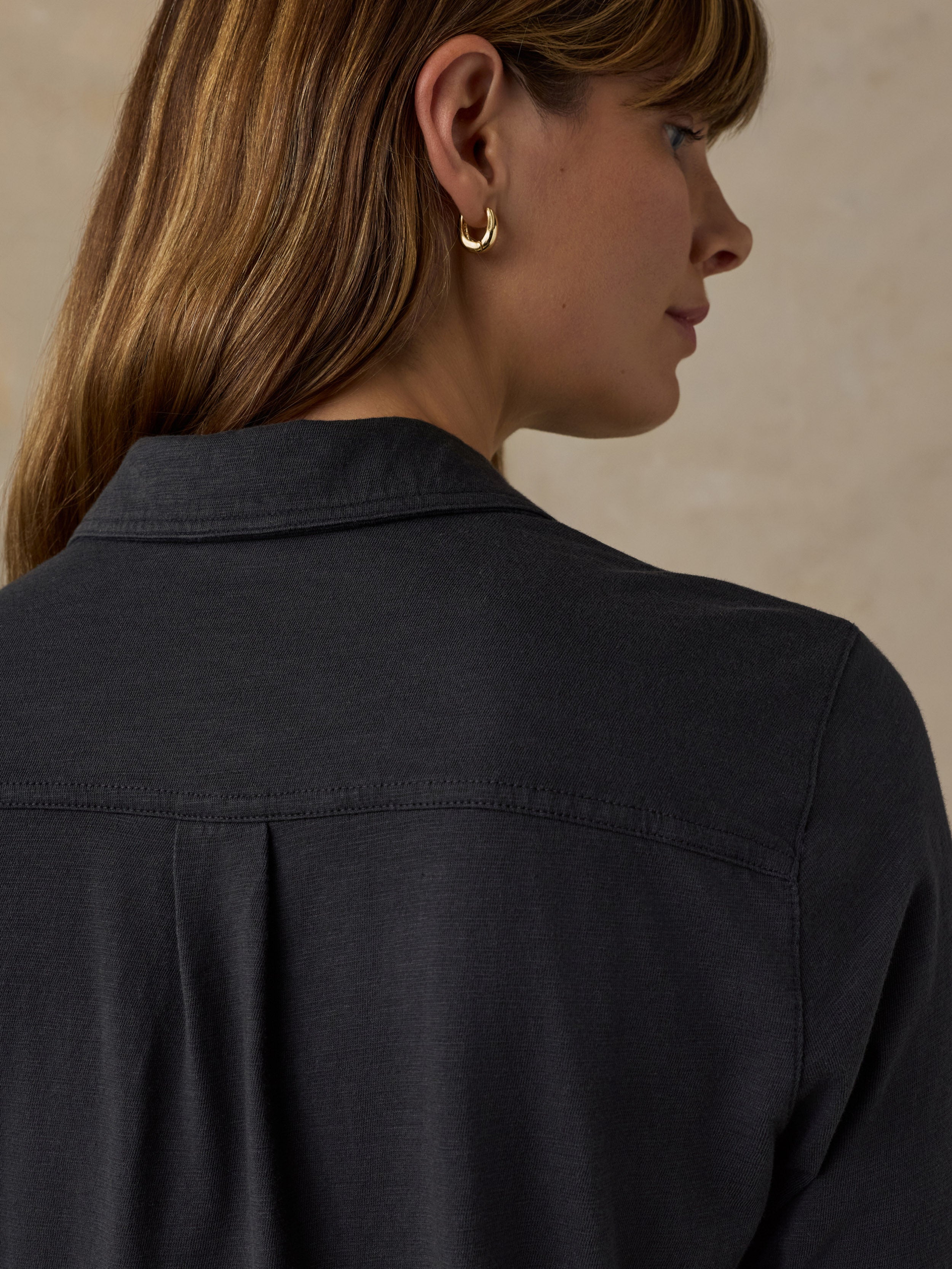 A close-up side profile of a woman wearing a dark charcoal gray button-up shirt and a small gold hoop earring, set against a neutral beige background.