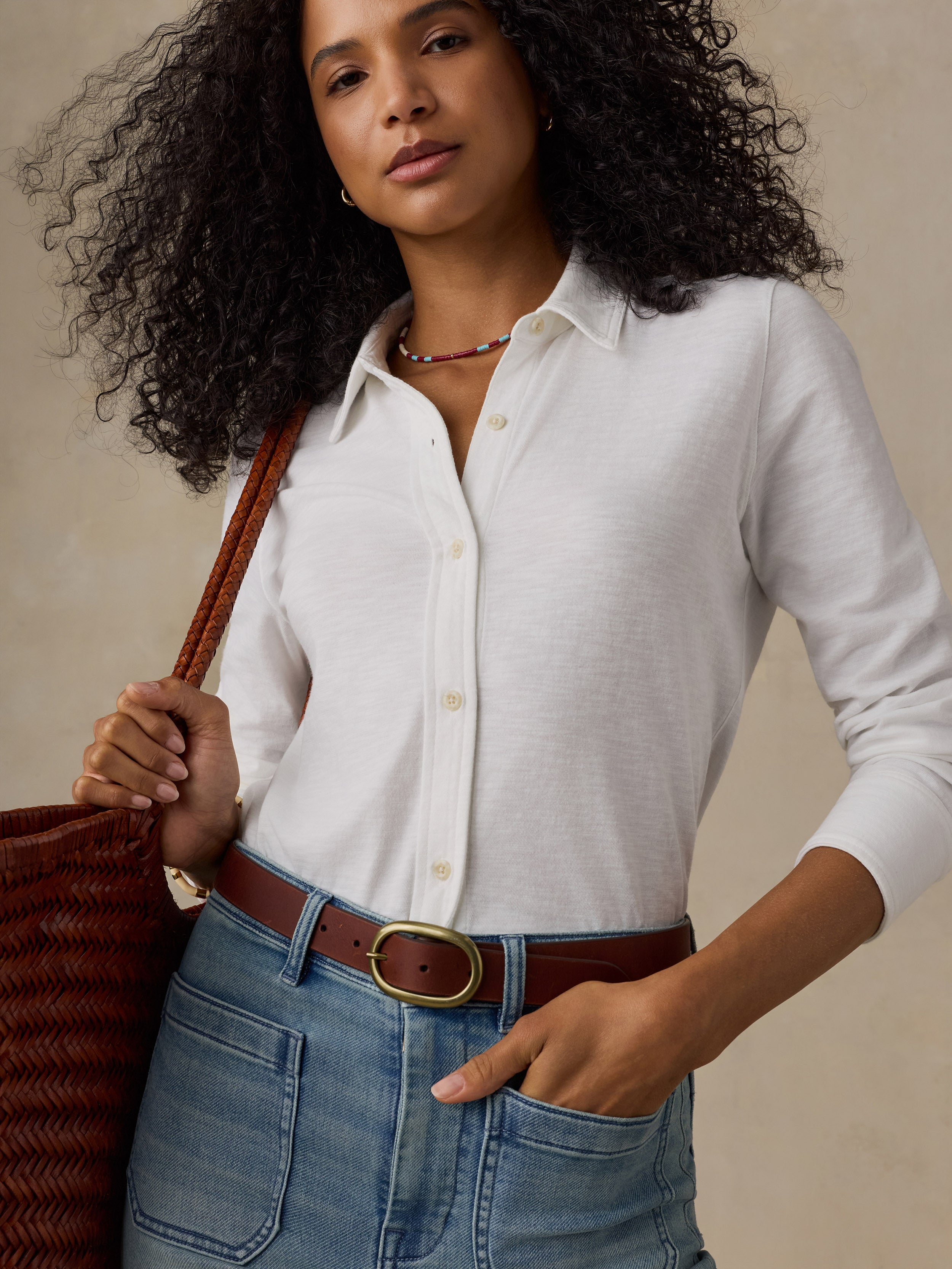 Woman wearing a white button down shirt with blue jeans and a woven bag against a beige background