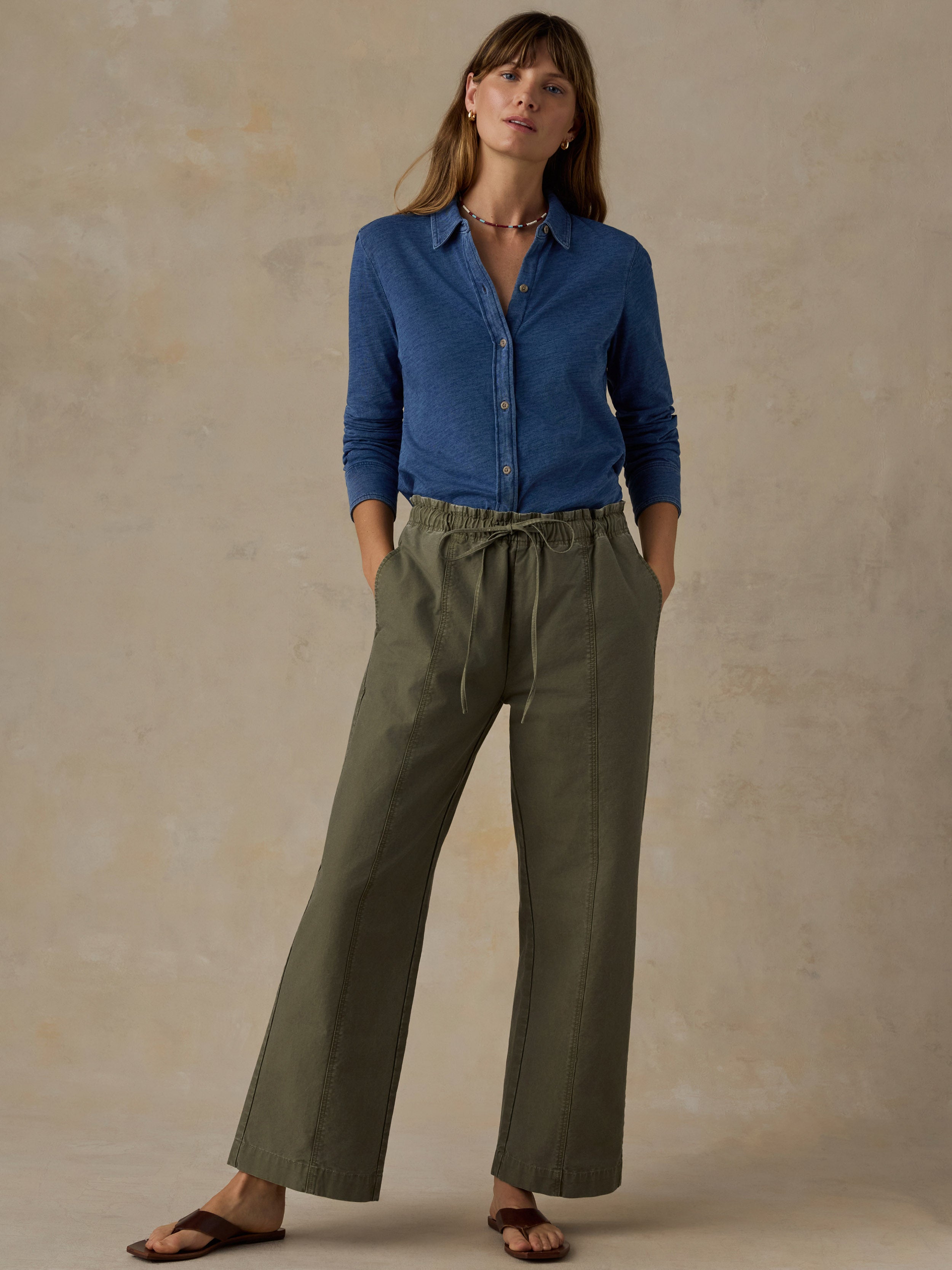 Woman wearing a blue button down shirt and olive green pants against a beige background