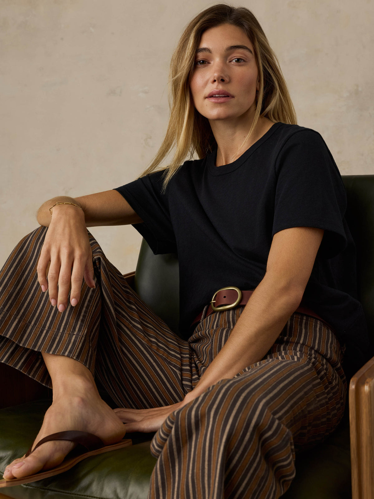 Woman sitting on a wooden chair wearing a black t-shirt and striped pants against a beige background, image 2 of 7