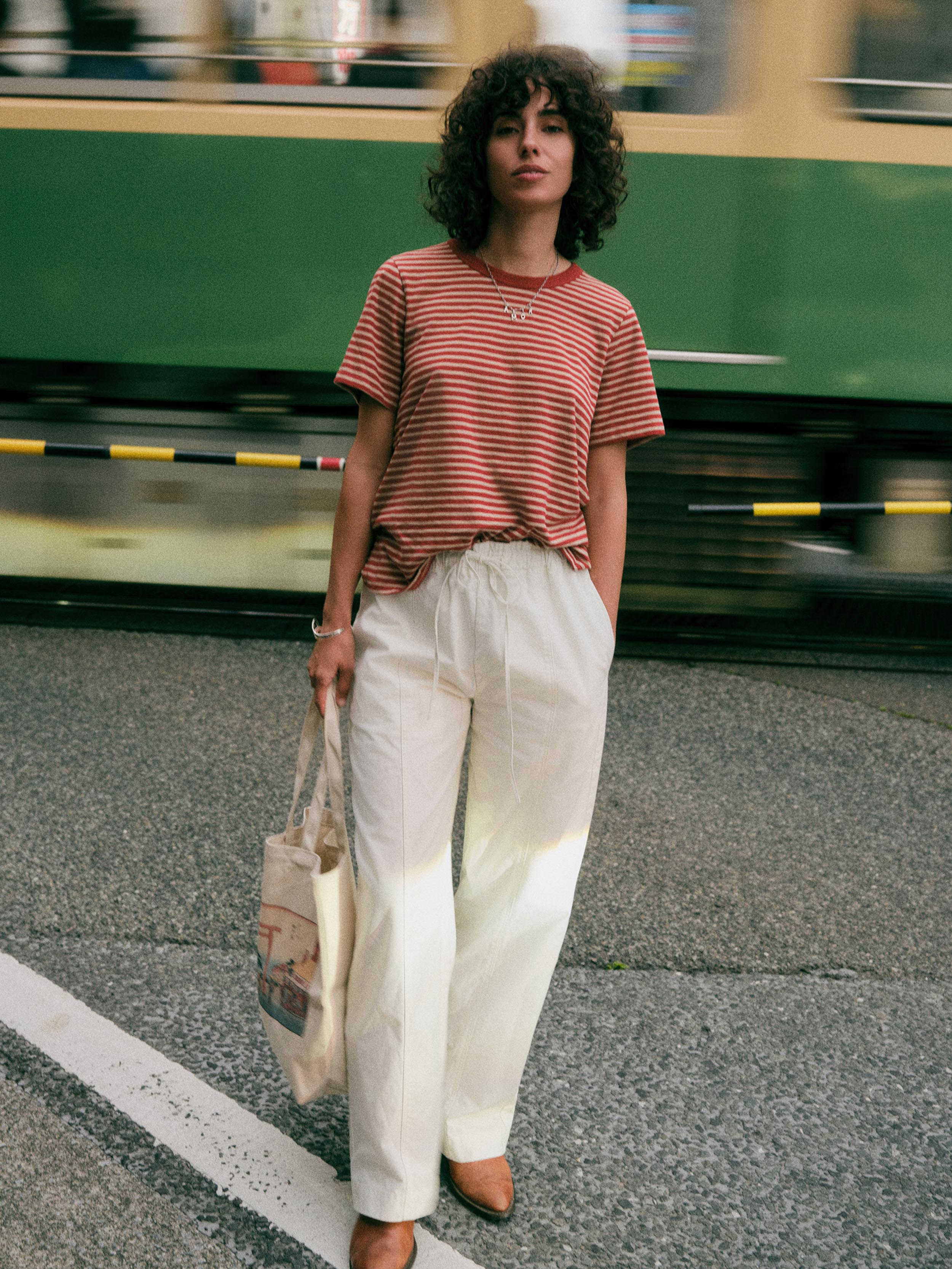 A woman stands on a street in front of a blurred green train, wearing a red and white striped short-sleeve t-shirt, cream wide-leg trousers, and brown shoes, while holding a canvas tote bag.