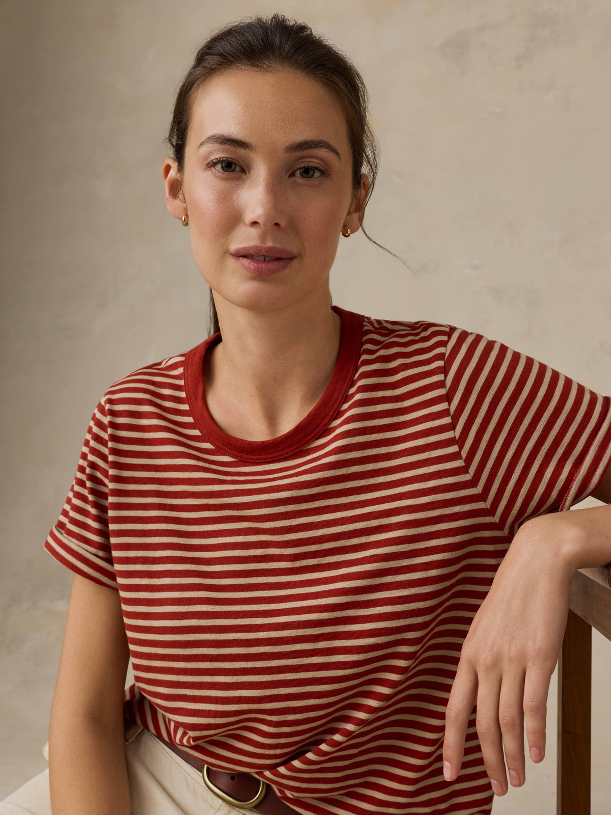 Woman sitting on a wooden chair wearing a red striped shirt against a beige background, image 6 of 8